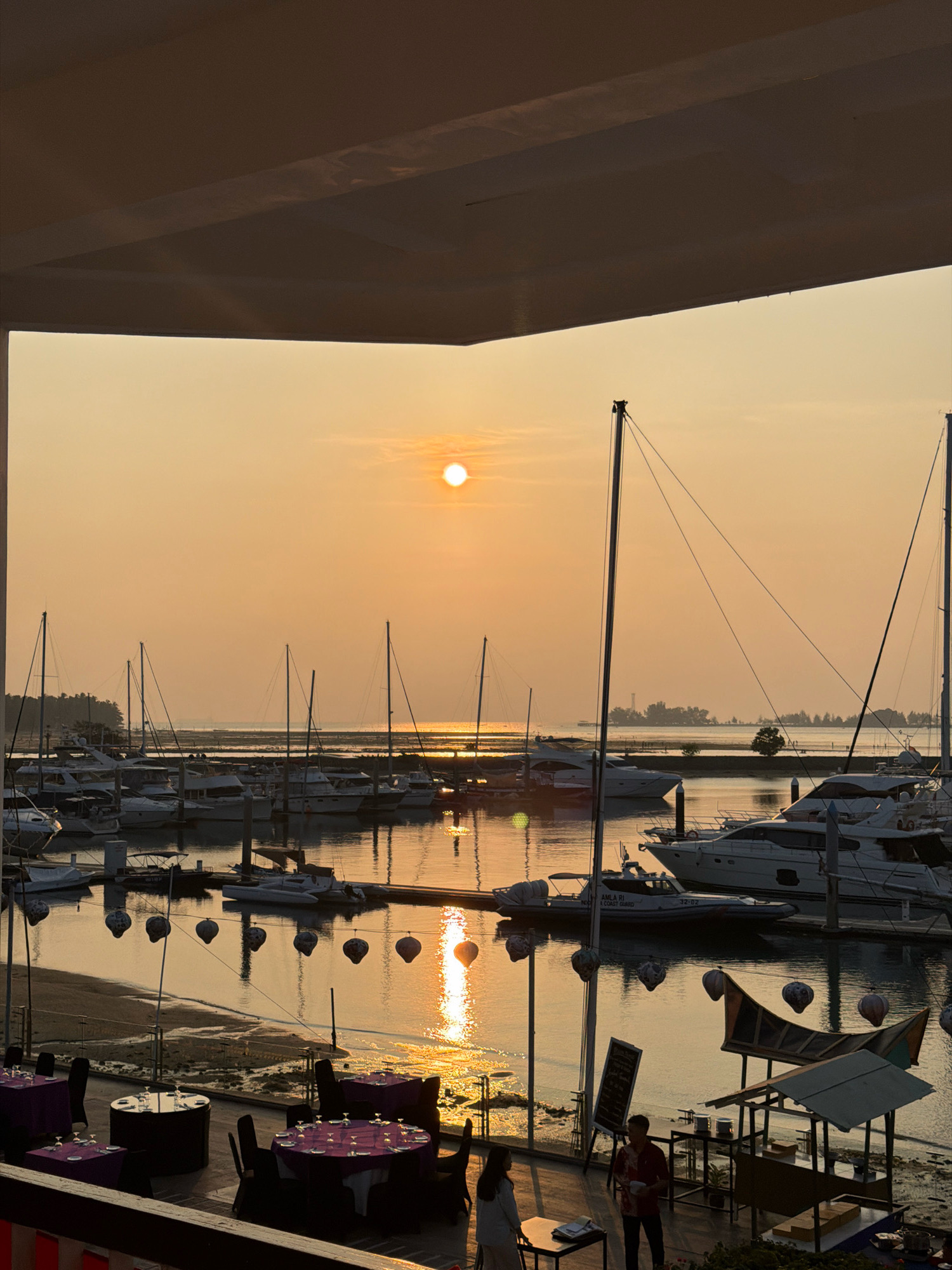 This image captures a serene sunset scene at a marina in Batam, Indonesia. The sky is awash in warm, golden hues as the sun descends towards the horizon. In the foreground, a covered outdoor dining area is set up with tables draped in purple cloths and set with glassware. Two individuals, a man and a woman, are conversing near a display board and a food stall. Beyond the dining area, a harbor is filled with various yachts and sailboats docked in the calm water. The reflections of the setting sun and the boats shimmer on the water's surface. In the distance, a hazy shoreline with trees and structures can be seen. Decorative paper lanterns are strung across the area, adding a festive touch to the tranquil evening. The overall impression is one of peacefulness and a beautiful natural spectacle.