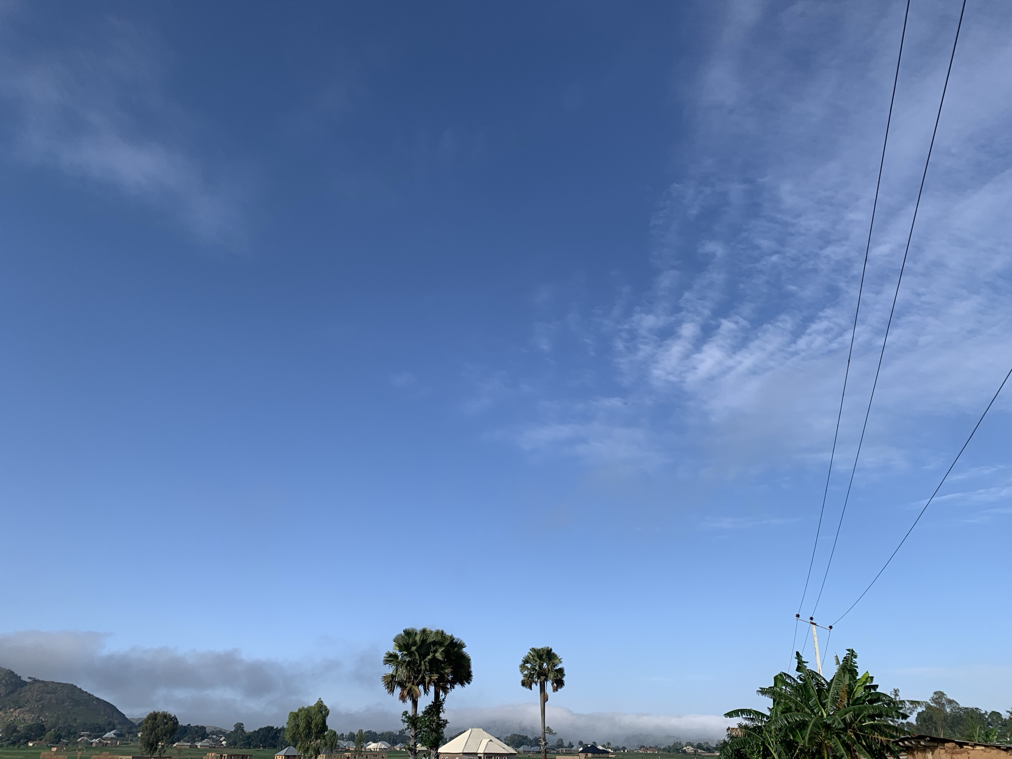This is a landscape photograph taken in Kuru Karama, Nigeria, featuring a clear blue sky with wispy clouds. In the foreground, several palm trees stand tall. To the right, a utility pole supports power lines that stretch across the frame. In the background, a small settlement with various buildings, including houses and what appears to be a mosque, is visible. A misty layer hangs low in the distance, partially obscuring a distant hill or mountain. The overall scene depicts a rural or semi-rural environment under a bright sky.