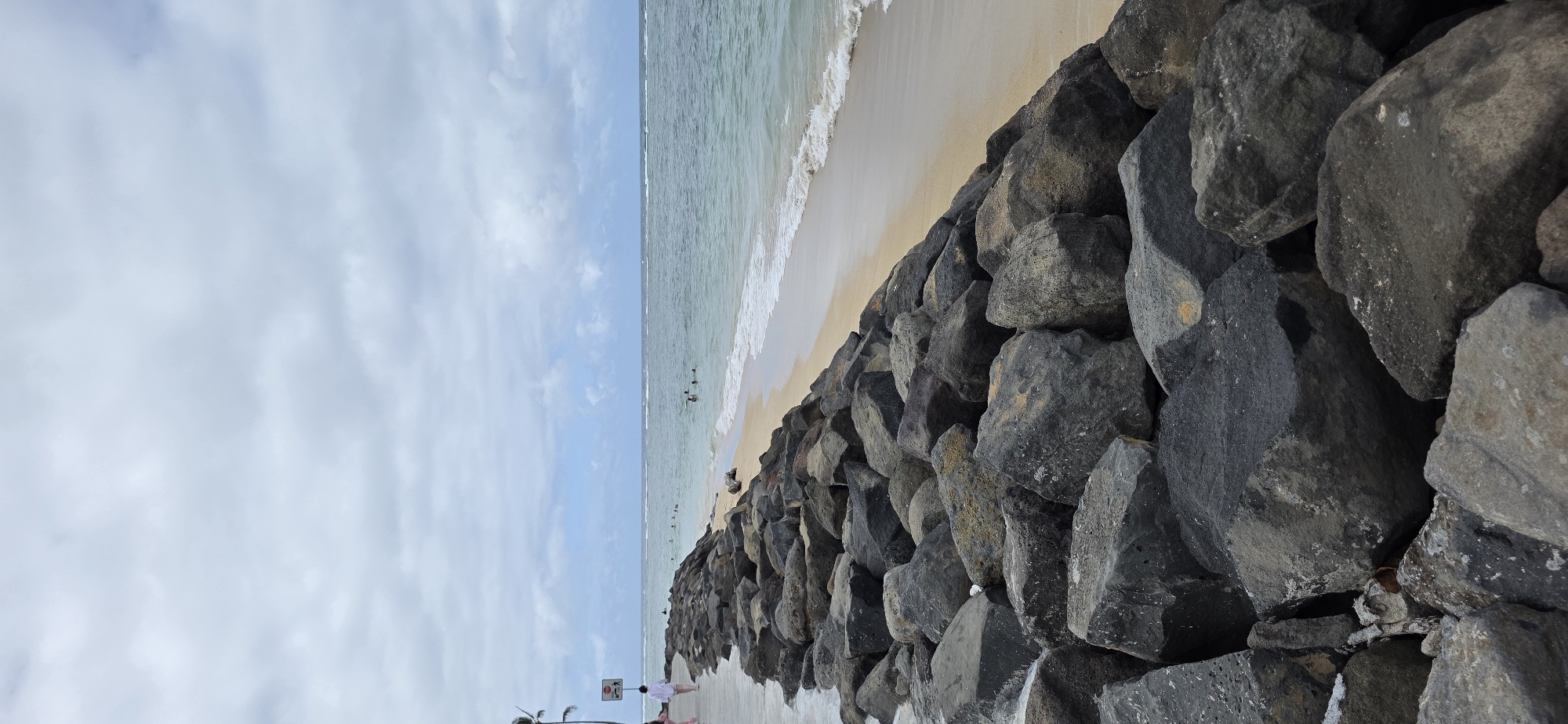 This is a full, vertical, outdoor, eye-level shot of a rocky shoreline in Kaaawa, Hawaii, United States of America. The foreground is dominated by a pile of large, dark gray and black rocks, some of which are partially submerged in the ocean. A light gray, concrete pathway runs along the top of the rocks on the left side of the image. A person in a white shirt and dark pants is walking on the pathway away from the camera. Further down the pathway, a stop sign with a stick figure walking is visible. The ocean stretches across the midground and background, with gentle waves lapping at a sandy beach on the right side of the image. Several people are visible swimming and wading in the ocean. The sky is overcast with thick, gray clouds, interspersed with patches of blue sky. A palm tree is partially visible on the far left.