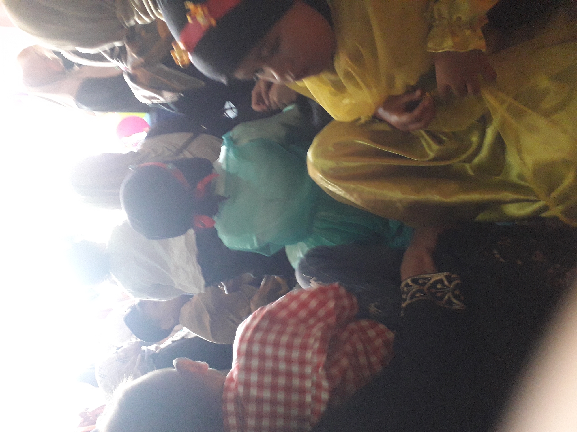 The image depicts a group of people, primarily children, gathered indoors. The lighting suggests it could be daytime with bright sunlight streaming in from the background, creating a blown-out effect in that area.
In the foreground, a child is visible from the shoulders up, facing left. They are wearing a black head covering with a red and yellow embellishment. To the left of this child, another child is seen from the back, wearing a red and white checkered shirt. This child's head is turned slightly to the left. Partially visible behind the child in the checkered shirt is a person in a black garment.
In the mid-ground, a child in a light blue garment is seated, facing away from the camera. To the left and slightly behind this child, a child in a tan outfit is also seated. Further in the background, several other individuals are present, some wearing headscarves. A pink and yellow balloon is visible in the far background, near a bright light source. The overall impression is of a casual gathering, possibly an event or celebration.
Without further information or context, the specific location within the city of Cankuang, Indonesia cannot be determined. However, the presence of individuals in what appear to be modest Islamic attire suggests a cultural setting where this is common. The gathering appears to be taking place in a room with a bright opening to the outside.