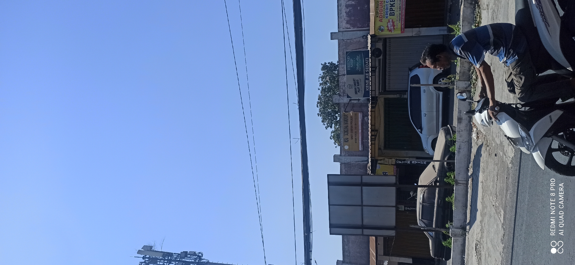 In Sedati Agung, Indonesia, under a clear blue sky, a man rides a white scooter down a street lined with buildings. The buildings are low-rise structures with faded facades, one displaying a sign for "KSU ADIGUNA MAKMUR" and another with "FRIENDSHIP Vodka Alia". A dark-colored sedan is parked on the side of the road, partially obscured by overgrown vegetation. A white minivan is also visible parked in front of one of the buildings. Power lines are strung across the sky. The foreground shows the rider and the front of the scooter. The overall scene depicts a typical street view in a local Indonesian town.