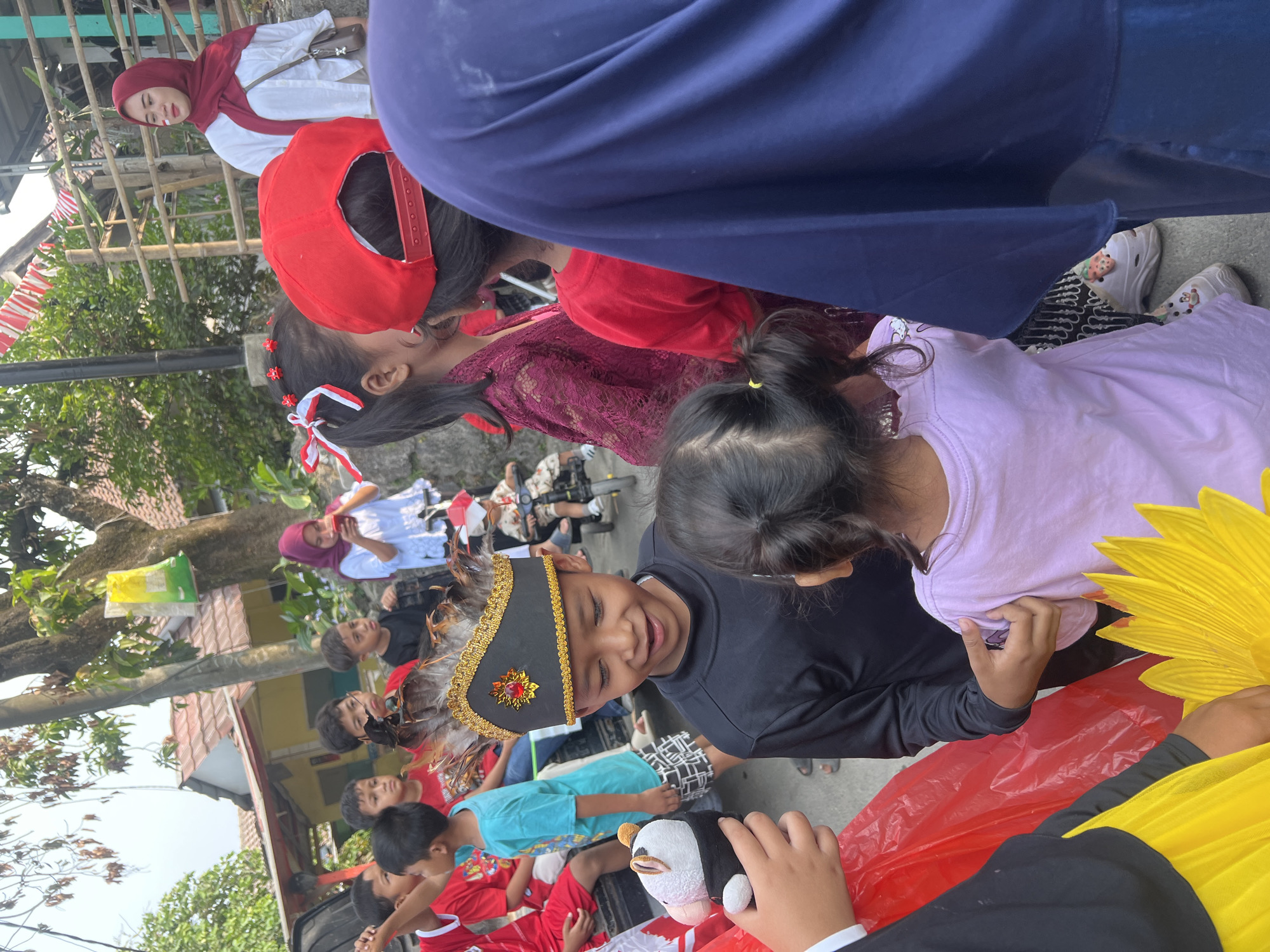 The image shows a group of children and adults gathered outdoors in what appears to be a residential area in Klapanunggal, Indonesia. In the foreground, a young boy wearing a traditional black headdress adorned with feathers and gold trim is looking down with a slight smile. He is interacting with a younger child wearing a lavender t-shirt. To the left, a child's hand holds a stuffed animal that resembles a white sheep wearing a black vest. Other children are visible in the background, some wearing red shirts. A woman in a red hijab is also present, holding a red cup. Decorative red and white banners are strung up in the background, suggesting a celebration or event. The environment includes houses with tiled roofs and trees.