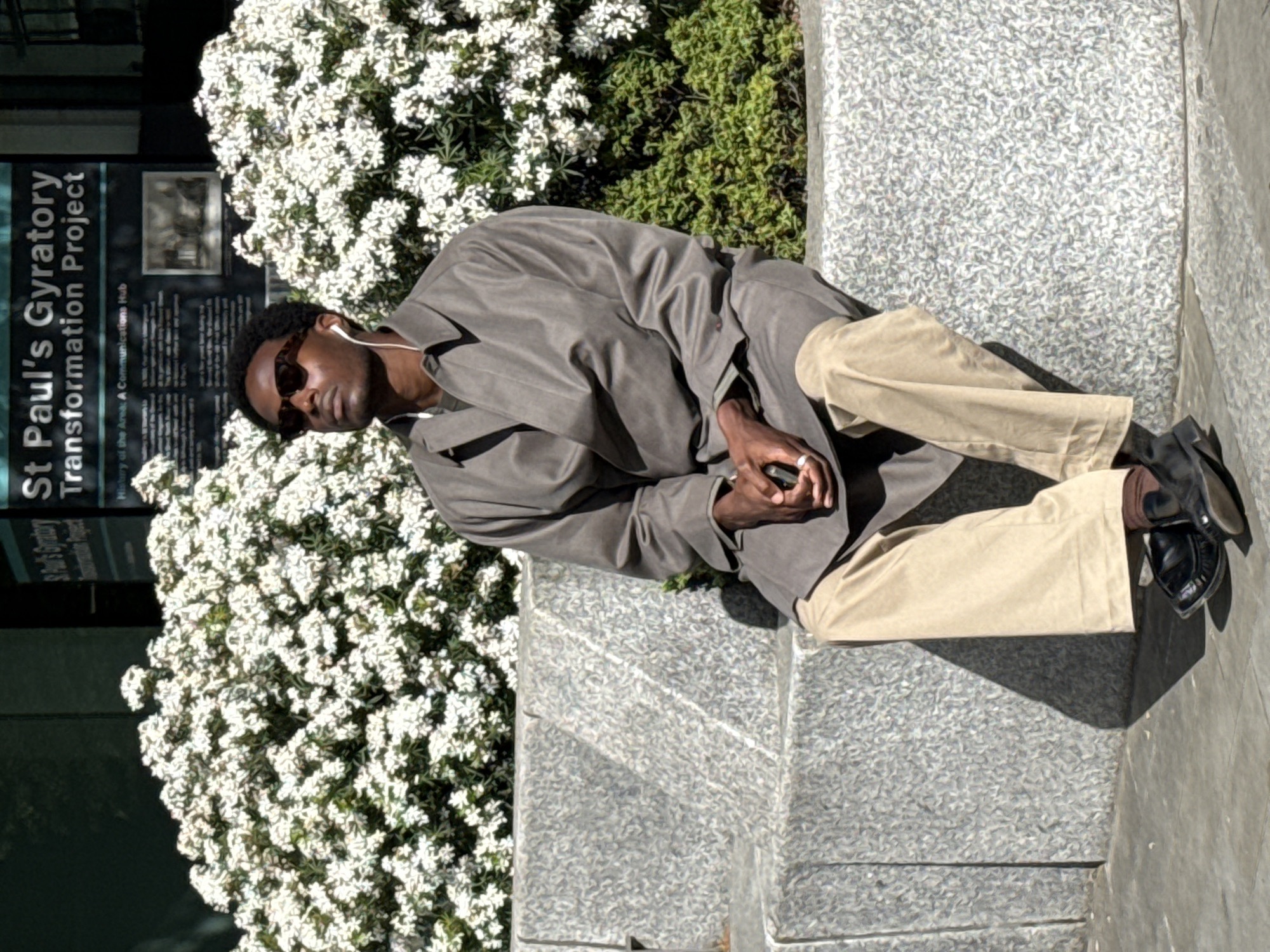 The image shows a person sitting on a concrete ledge next to a sign that reads "St Paul's Gyratory Transformation Project," suggesting a location in the city of London, United Kingdom. The person is wearing sunglasses, an earbud, a gray jacket, and tan pants, and they are holding a phone in their hands. The person is positioned in front of white flowering bushes. The location appears to be outdoors on a sunny day.