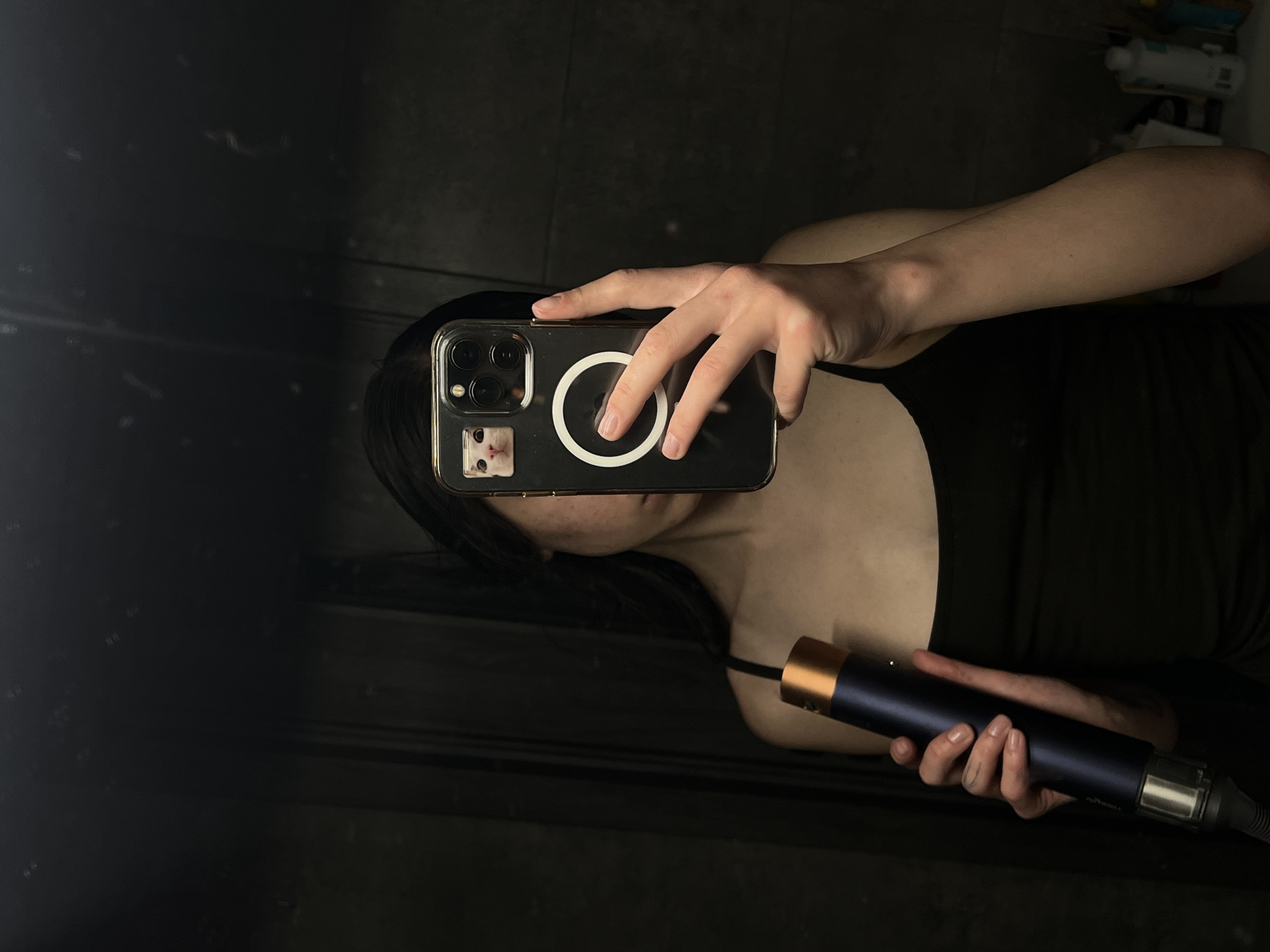 In a dimly lit room with dark, textured walls, a person with long dark hair is holding a dark blue cylindrical device with a gold top in their left hand. They are taking a mirror selfie with a smartphone held in their right hand. The smartphone has a clear case with a small sticker of a cat's face in the top left corner, and a white circular magnetic accessory on the back. The person is wearing a black tank top. The room appears to be a bathroom, with reflections of what might be tiles or other bathroom fixtures visible in the mirror. There is no indication of the specific location within Ternopil, Ukraine.