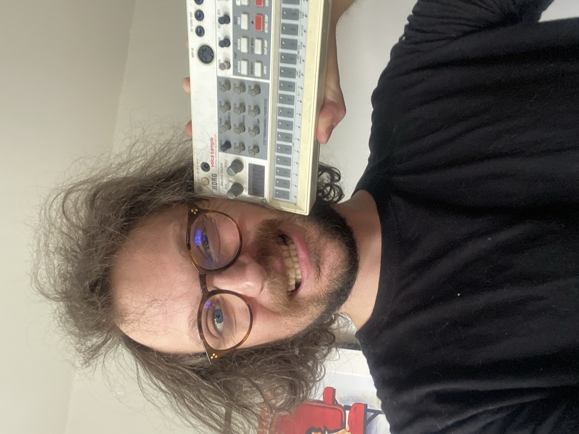 A man with long, curly brown hair and a beard is holding a Korg Volca Sample digital sample sequencer up to his face. He is wearing glasses with brown frames and a black t-shirt. He is smiling, and his teeth are visible. The background is a plain white wall. Part of a picture can be seen in the bottom left corner. The image is oriented vertically. There is no indication of the location being Rusanovo, Russia.