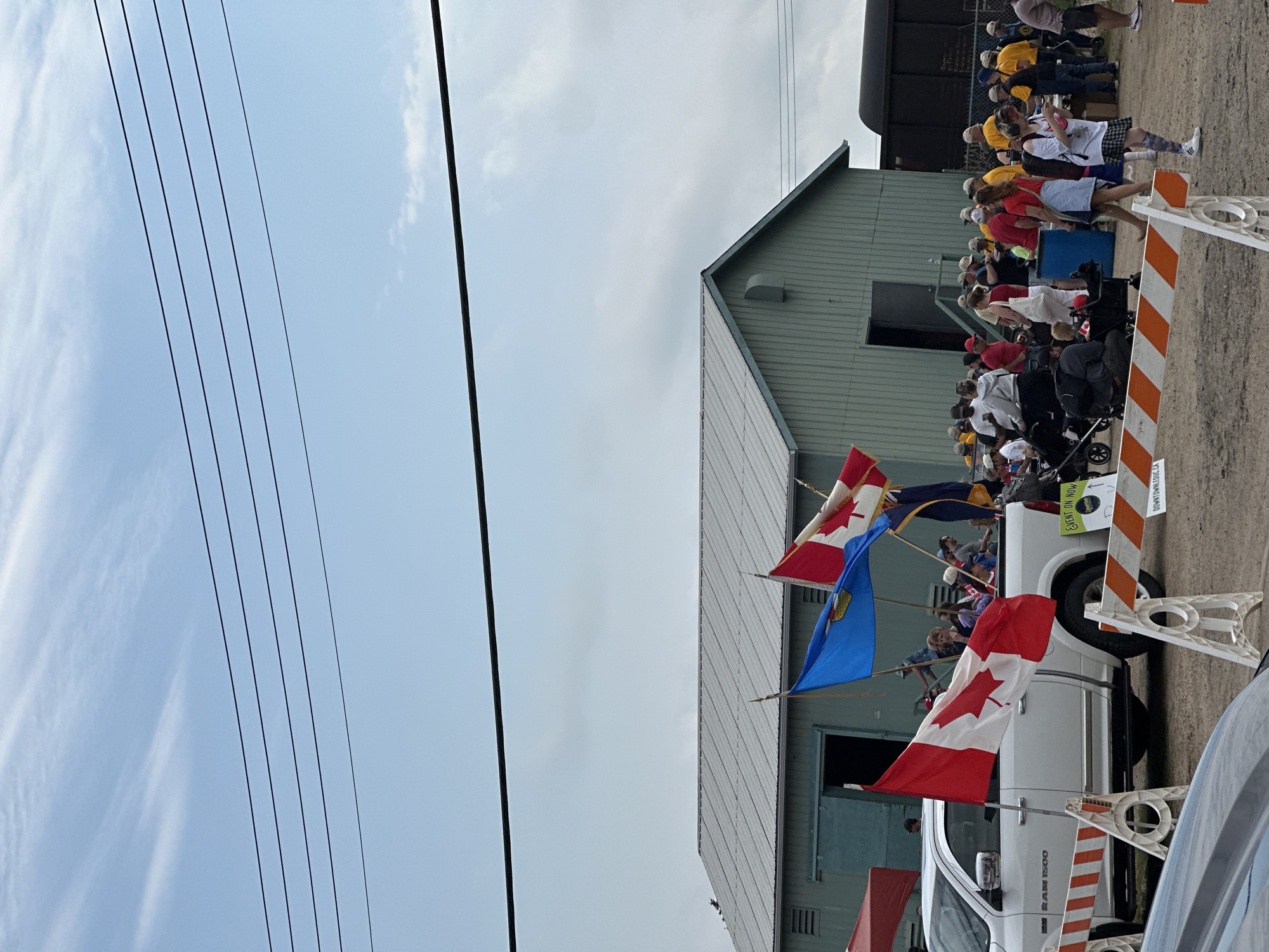 The image depicts an outdoor scene with a group of people gathered, many appearing to be spectators. A white pickup truck, adorned with Canadian flags, is the central object. Several people are standing or sitting in and around the truck. Some are holding flags and appear to be part of a procession or event. The truck has "RAM 1500" visible on its side.
In the background, a light-colored building with a sloped roof is visible, and further back, a dark train car or structure. The ground appears to be dirt or gravel. Orange and white safety barriers are placed near the truck. Overhead, power lines stretch across the frame against a bright, cloudy sky. The weather appears to be overcast or partly cloudy.
The presence of Canadian flags suggests a Canadian event, and the "City of Leduc, Canada" context places the scene specifically in Leduc. The people's demeanor is difficult to ascertain fully due to the image orientation and distance, but the overall atmosphere seems to be one of a community gathering or celebration. There is text visible on a sign attached to the truck that reads "EVENT ON NOW" and "DOWNTOWNLEDUC.CA". The overall impression is of a parade or festival.