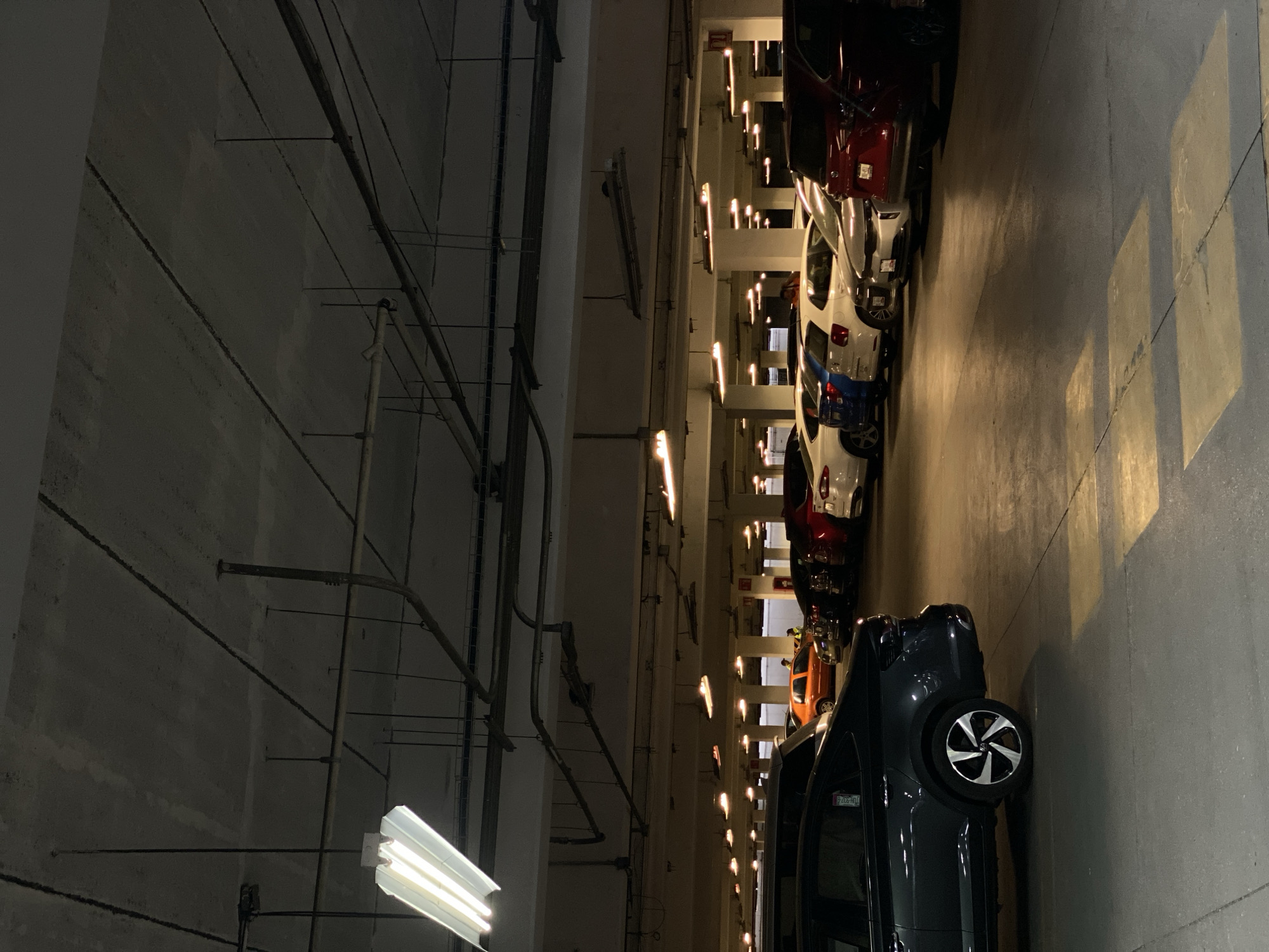The image shows an indoor parking garage in San Bernardino Tlaxcalancingo, Mexico. A row of parked cars of different colors and models lines the right side of the frame. The left side features a concrete wall with exposed pipes and wiring, illuminated by a fluorescent light fixture. The garage's interior is lit by overhead lights that create patterns on the concrete floor. No people are visible.