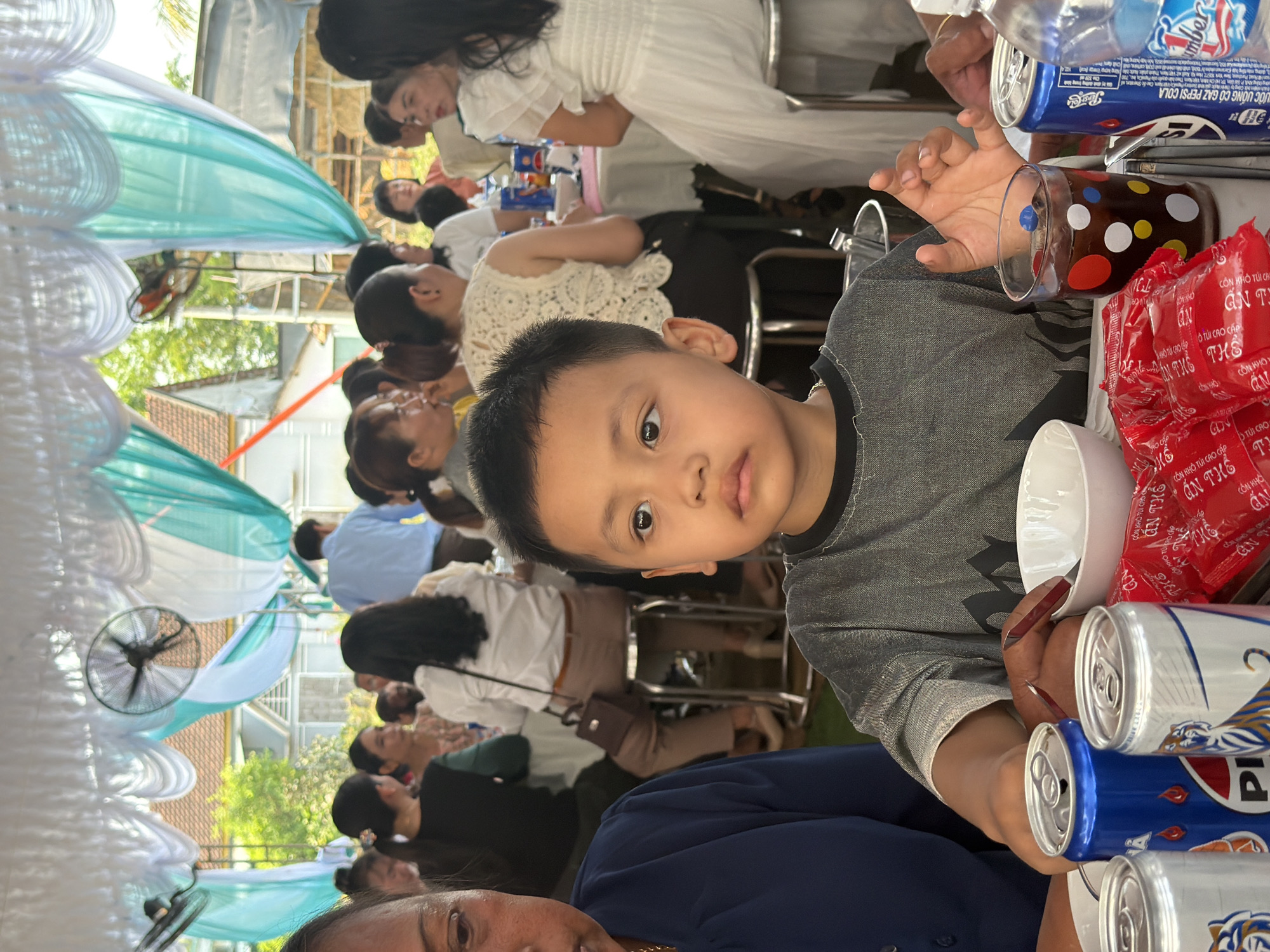 A horizontal medium shot depicts a young boy holding a clear polka-dotted glass, a white bowl, and several beer cans, seated at a table amid a gathering of people in what appears to be Bình An, Vietnam. The boy, with dark hair and light skin, looks directly at the camera. The table is covered with red snack packages and blue soda cans.
The background shows a crowded outdoor event with many people sitting at tables. The tables are decorated with turquoise and white draping, and there are fans attached to the structure above. The environment suggests a celebratory occasion, possibly a wedding or a large family gathering. The location is indicated as being in Bình An, Vietnam.