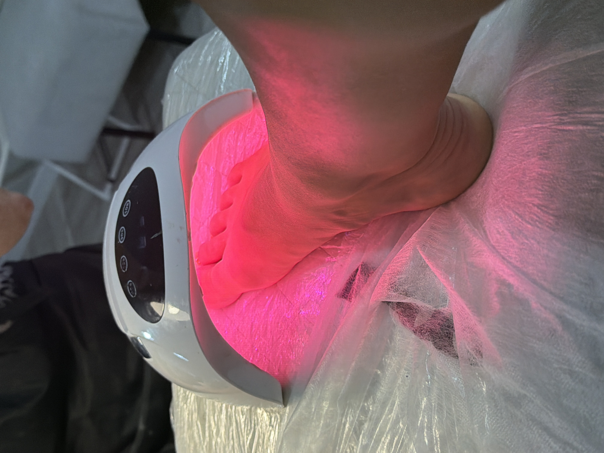 In Sochi, Russia, a person's foot is placed inside a white UV nail lamp that emits pink light. The lamp is positioned on a surface covered with white paper, possibly for hygiene. The background is blurred, but suggests an indoor setting, possibly a nail salon. The image captures a moment during a pedicure or nail treatment.
