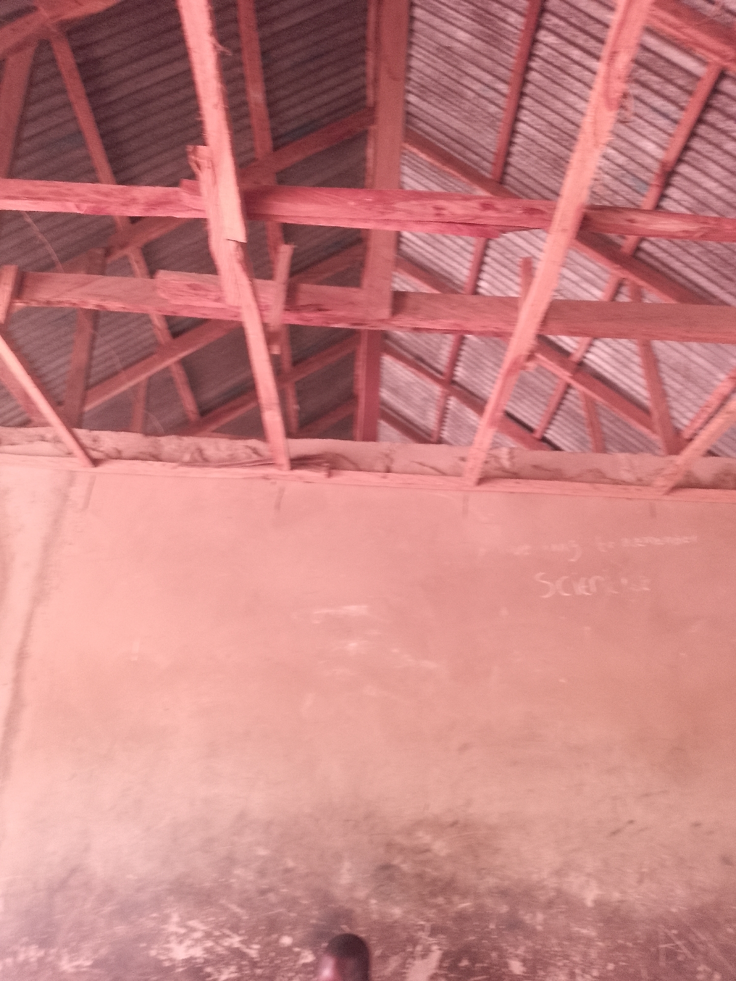 This media file depicts the interior of a building under construction or in disrepair, focusing on the roof structure and a section of wall. The roof is a wooden frame with corrugated metal sheeting. The wooden beams are rough-hewn and appear unfinished. The wall below is a pale, textured surface, possibly plaster or stucco, with some visible staining or wear.
On the wall, there is faint writing in what appears to be chalk. The word "Science" is clearly legible, written in large, somewhat stylized letters. Above it, there is some fainter text that is difficult to decipher, but it seems to be a phrase or sentence.
The overall impression is of a simple, possibly rural, structure. The lighting suggests it is daytime, but the exact time of day is not discernible. There are no people fully visible, although a dark, indistinct shape at the bottom of the frame might be a person's shoulder or head. The scene conveys a sense of utility and possibly neglect, with the focus on the building's construction rather than any specific activity. The red tint in the image might be due to the lighting conditions or an artifact of the camera.