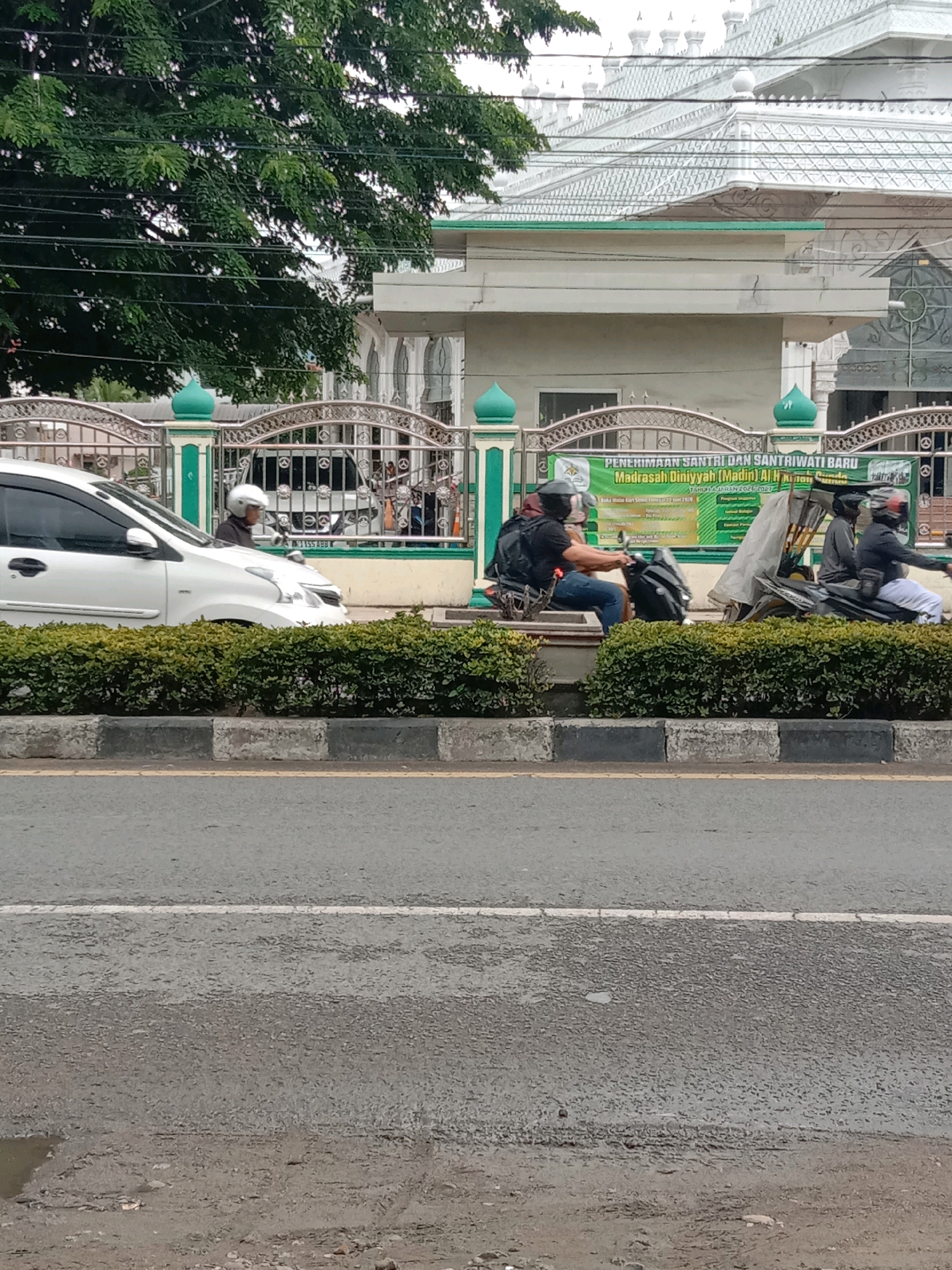 The image shows a street scene in Lhokseumawe, Indonesia, with a white car on the left and two motorcycles with riders on the right. A large tree with dense foliage is visible on the left side, partially obscuring a building with a distinctive white, multi-domed roof and green accents, which appears to be a mosque or religious institution. A decorative metal fence with green posts runs in front of the building. A green banner with text in Indonesian, including "PENERIMAAN SANTRI DAN SANTRIWATI BARU MADRASAH DINIYYAH," is displayed behind the fence. The road in the foreground has a striped curb and a white line indicating a lane divider. The ground to the right of the road appears to be unpaved and shows signs of wear and tear, including a small puddle of water. The sky is overcast, suggesting a cloudy day.