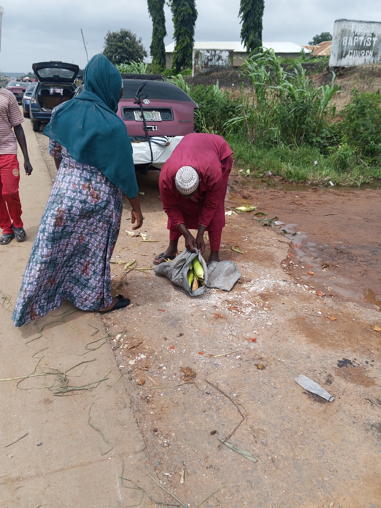 In the city of Oduape, Nigeria, an outdoor scene captures individuals interacting with produce near a roadside. The ground is a mix of paved surfaces and dirt, with scattered debris and a small puddle of water. In the background, vehicles are visible, suggesting a public area with traffic.
One woman, wearing a teal headscarf and a patterned skirt, stands with her back to the camera, her gaze directed towards the activity unfolding. To her right, a man in a maroon outfit and a white cap is bending down, tending to a grey sack filled with what appears to be corn. He is in the process of sorting or inspecting the produce.
To the left of the woman, a person wearing a striped shirt and red pants is also present, though their interaction with the produce is less clear. They are facing away from the viewer, with their left hand slightly raised.
The environment shows signs of natural elements, with tall green plants and grass growing along the roadside. A white sign with black lettering, partially obscured by vegetation, reads "FIRST BAPTIST CHURCH," indicating a nearby religious institution. The overall impression is a snapshot of daily life and commerce taking place in a public space within Oduape.