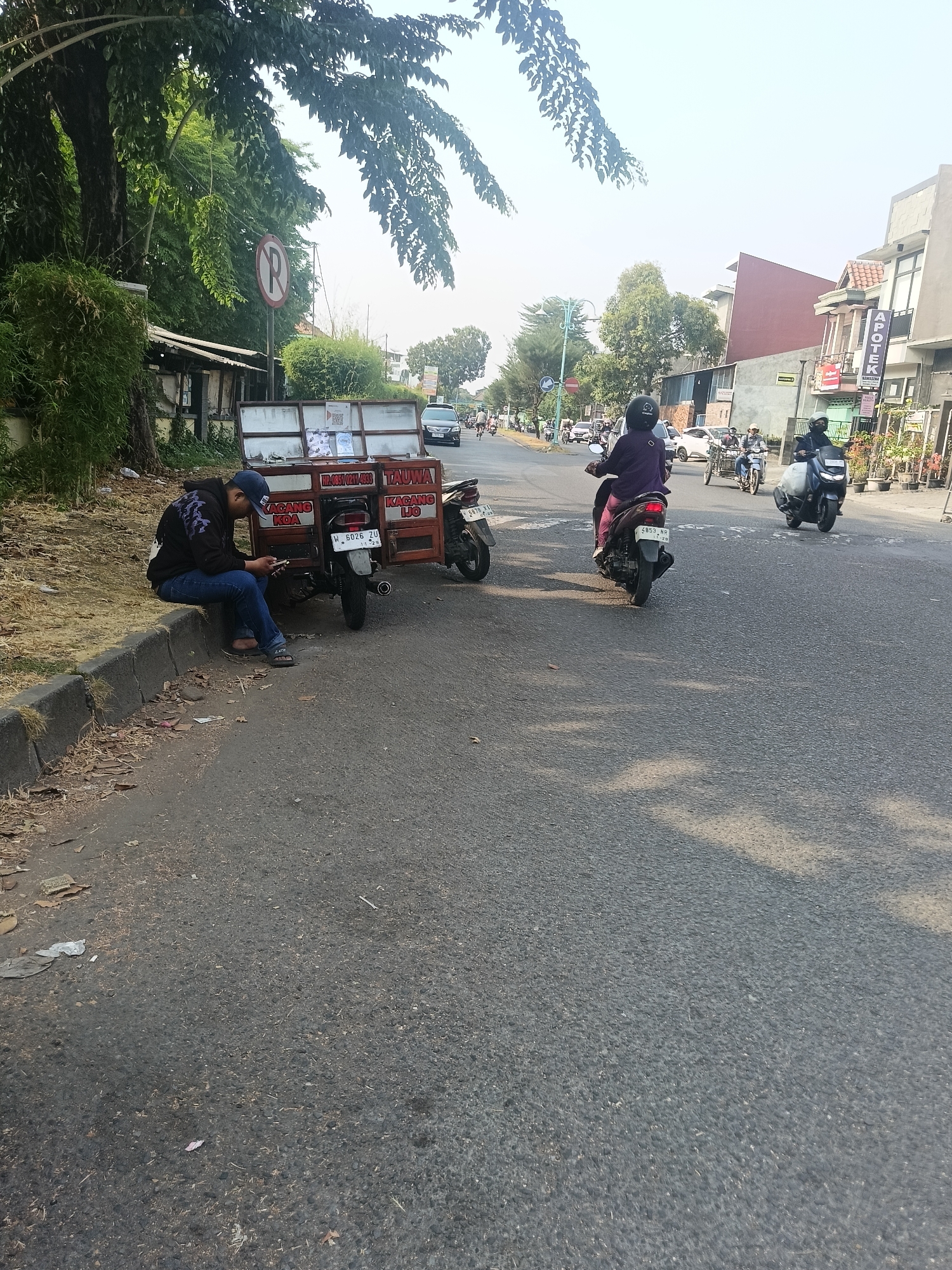 The image depicts a street scene in Wedi, Indonesia. In the foreground, a young man wearing a blue baseball cap, dark hoodie with a purple design, and blue jeans sits on a curb next to a small, three-wheeled motorcycle cart. He is looking down at his mobile phone. The cart is orange and has various signs attached, including one that reads "KACANG KOA" and another with Indonesian text. Behind the cart, the street is paved, and several other motorcycles are visible, some with riders. In the mid-ground, a woman on a purple motorcycle rides away from the camera, and further down the street, other vehicles and buildings line the road. The buildings appear to be shops or residences, with one displaying a sign for "APOTEK" (pharmacy). Trees and foliage are present on the left side of the image and further down the street. The sky is clear and bright, indicating daytime. A "no parking" sign is visible on a pole to the left.