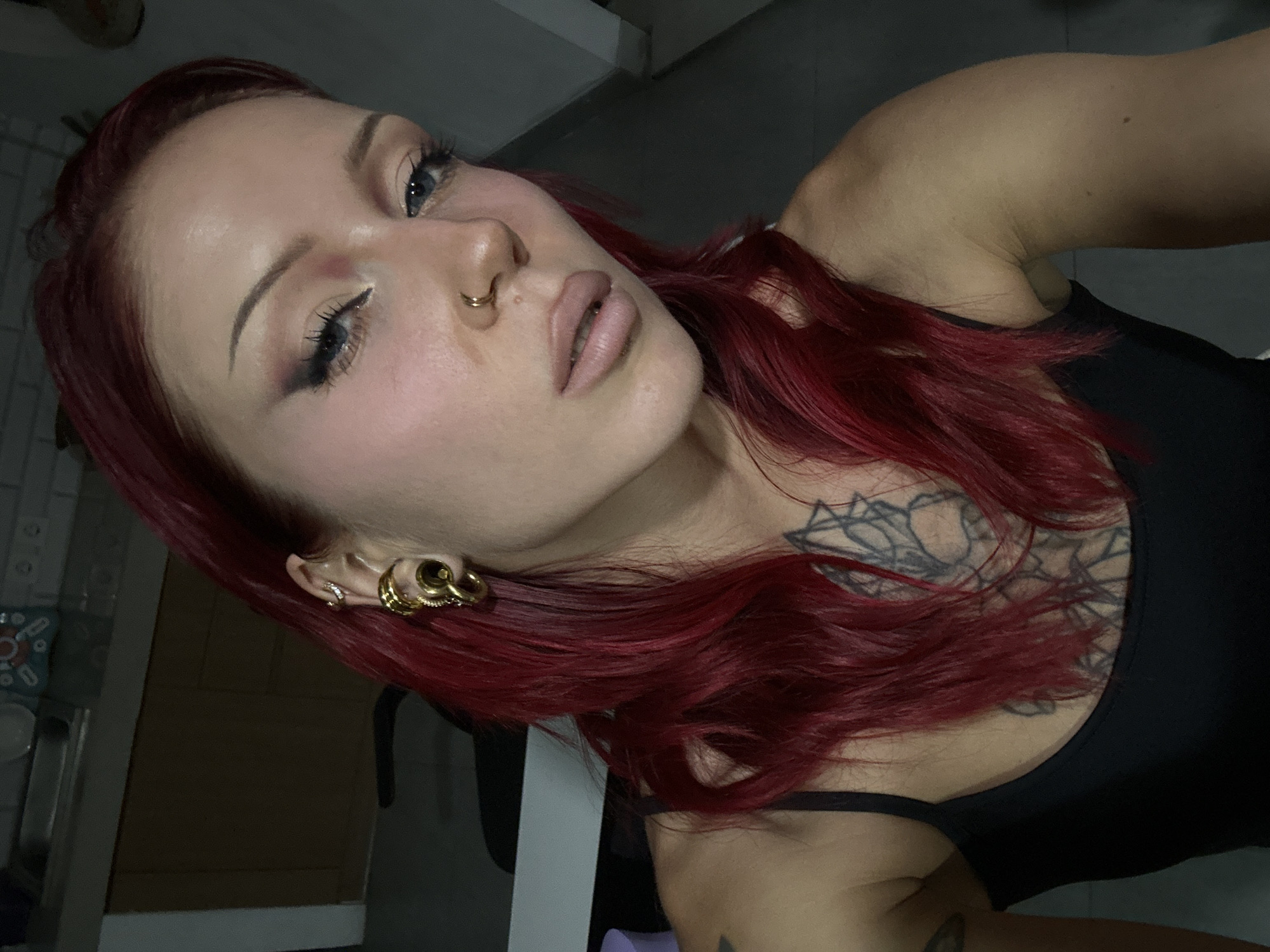 A young woman with vibrant red hair and striking makeup is captured in a close-up, slightly angled selfie. She wears a black tank top, revealing a portion of a dark, intricate tattoo on her chest and shoulder. Her left ear is adorned with multiple gold piercings, including a large, spiraled earring. She also has a gold hoop piercing in her nose. Her eyes, with dramatic winged eyeliner and eyeshadow, are looking slightly off-camera. Her lips are painted a neutral matte shade. The background appears to be an indoor setting, possibly a kitchen or living area, with a countertop and cabinets visible in the upper left corner. The lighting is dim, creating shadows that highlight her features. The overall impression is one of self-expression and bold personal style.