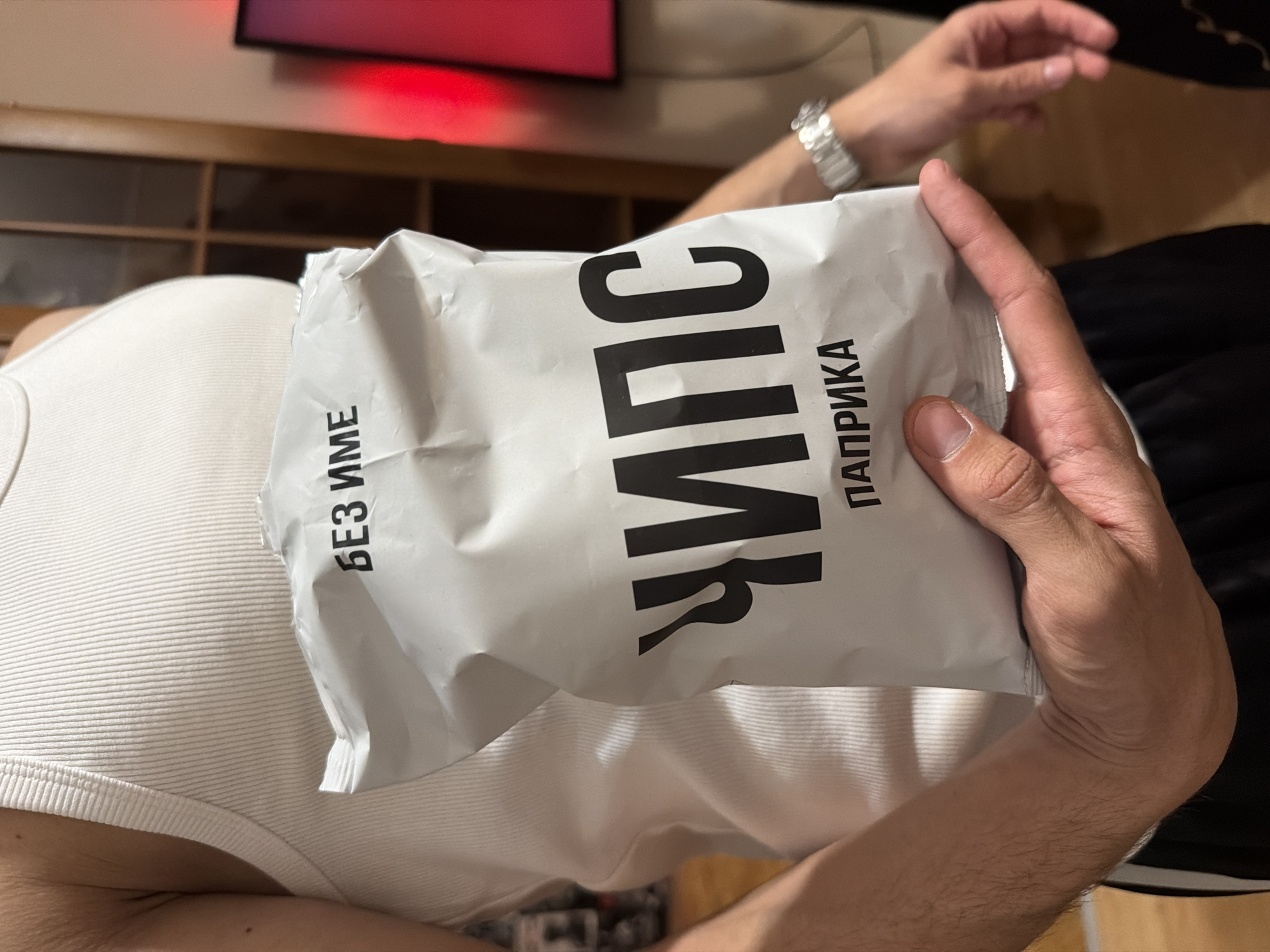A man holds a white bag of "CHIPS" paprika flavored chips on his chest while sitting indoors. The text on the bag is in Russian. The location is unknown, but the brand of chips is a brand that is sold in Sofia, Bulgaria. The man wears a white ribbed tank top and a silver wristwatch. The background includes a wooden cabinet, a television with a red glow, and the man's other arm with a black and white striped sleeve.