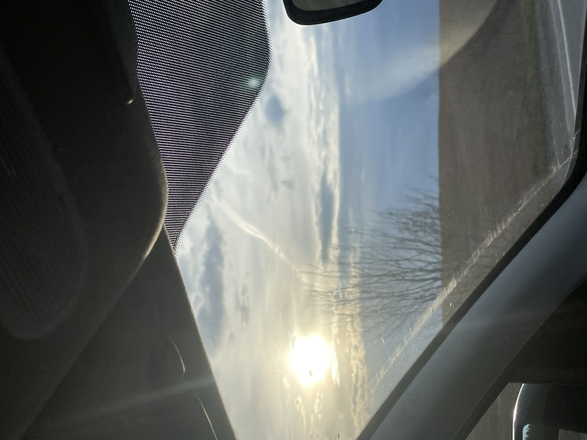 The image presents a view looking upwards through the panoramic roof or windshield of a vehicle, capturing a dynamic sky with the sun prominently featured. The perspective is from inside the car, directed towards the sky and reflections on the glass.
The sky above is a blend of clear blue patches and scattered white clouds. A brilliant, low-lying sun creates a strong glare and lens flare in the lower part of the frame. A faint, white contrail from an aircraft is visible cutting across the cloudy sky. Through the glass, the intricate, bare branches of a tree, indicative of winter or early spring, are discernible on the right side, partially masked by the sun's intensity and reflections. Further to the right, a dark, solid structure, possibly a building or a wall, is seen through the glass.
Elements of the car's interior frame the scene. On the left, a dark, textured surface, likely a part of the headliner or an interior trim, is visible. The edges of the glass, particularly the top-left and left sides, display a distinctive black frit band pattern, which consists of small dots. Minor streaks and smudges are present on the glass surface.
The low angle of the sun suggests the time of day is either early morning or late afternoon. The scene conveys a sense of being in transit or stationary within a vehicle, under a sunny and partly cloudy sky. The described location is Povarenki, Russia. No discernible text is visible within the image.
