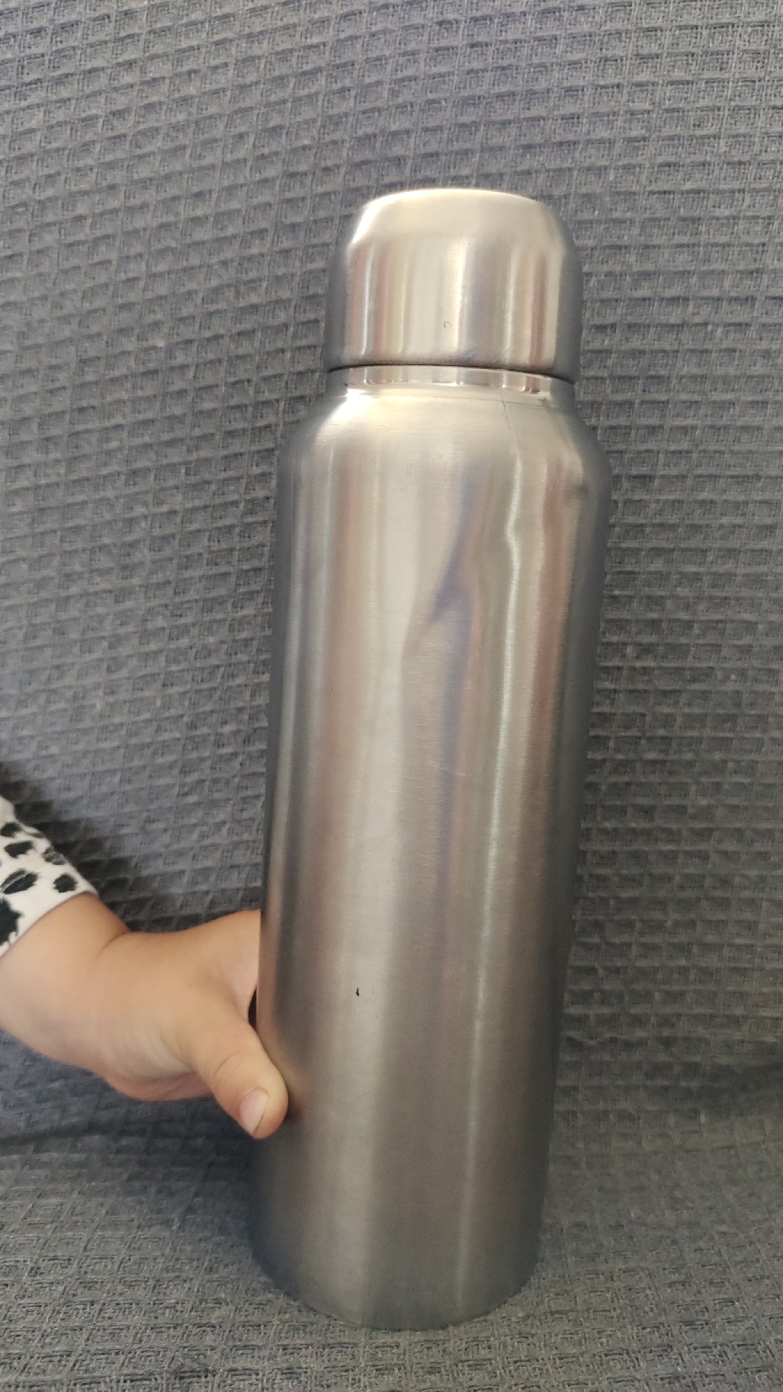 An aluminum water bottle stands upright on a gray textured surface, with a child's hand in the lower left corner grasping its lower portion. The bottle is cylindrical with a rounded top, appearing to be made of brushed metal, reflecting light from above and the side. The surface beneath is a woven fabric with a repeating diamond pattern. The background is a solid gray, textured wall. The image is well-lit, with no visible shadows indicating a specific time of day or artificial light source. The location is Unknown, Unknown.