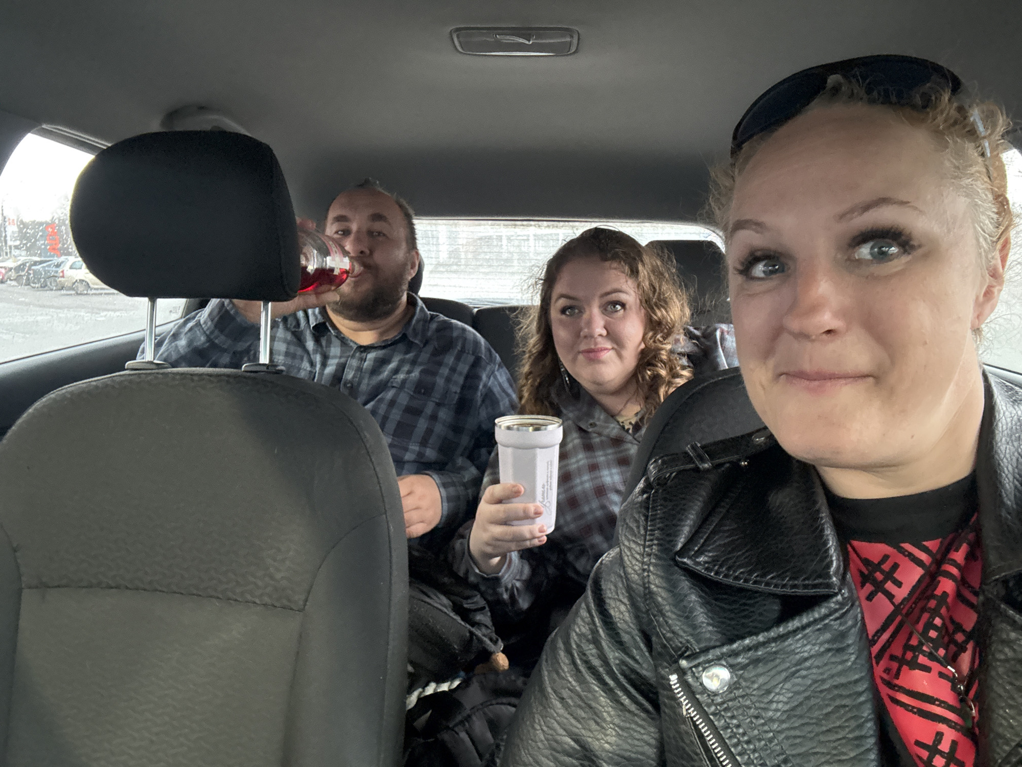 Three individuals are inside a car. In the back, a man drinks from a clear bottle with a red liquid, while a woman holds a white tumbler. The person in the front is likely taking a selfie. Outside, it appears to be raining or wet, and there's a parking lot with vehicles, as well as a building in the distance. The location is Izhevsk, Russia.