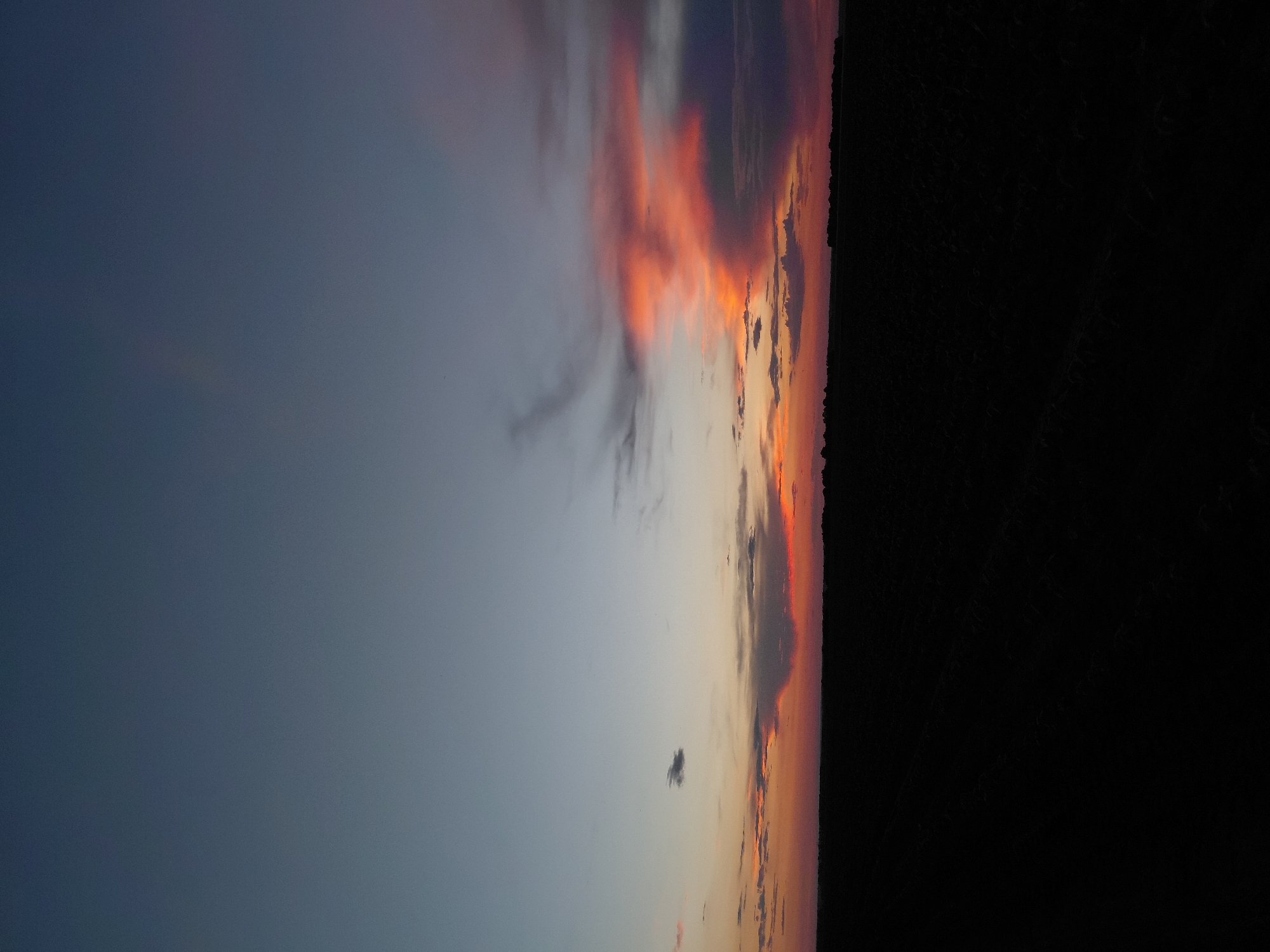 The image shows a twilight sky over a dark, silhouetted field. The sky is a gradient of colors, transitioning from a deep blue at the top to lighter shades of orange and pink near the horizon. Scattered clouds catch the last rays of sunlight, creating vibrant streaks of red and orange. The foreground is a flat, dark expanse, likely a tilled field, with faint patterns suggesting rows. The horizon line is sharp and distinct, with a line of trees barely visible in the distance. There are no people or specific objects visible in the image, making it a general landscape scene. The location is not specified in the image or its description.