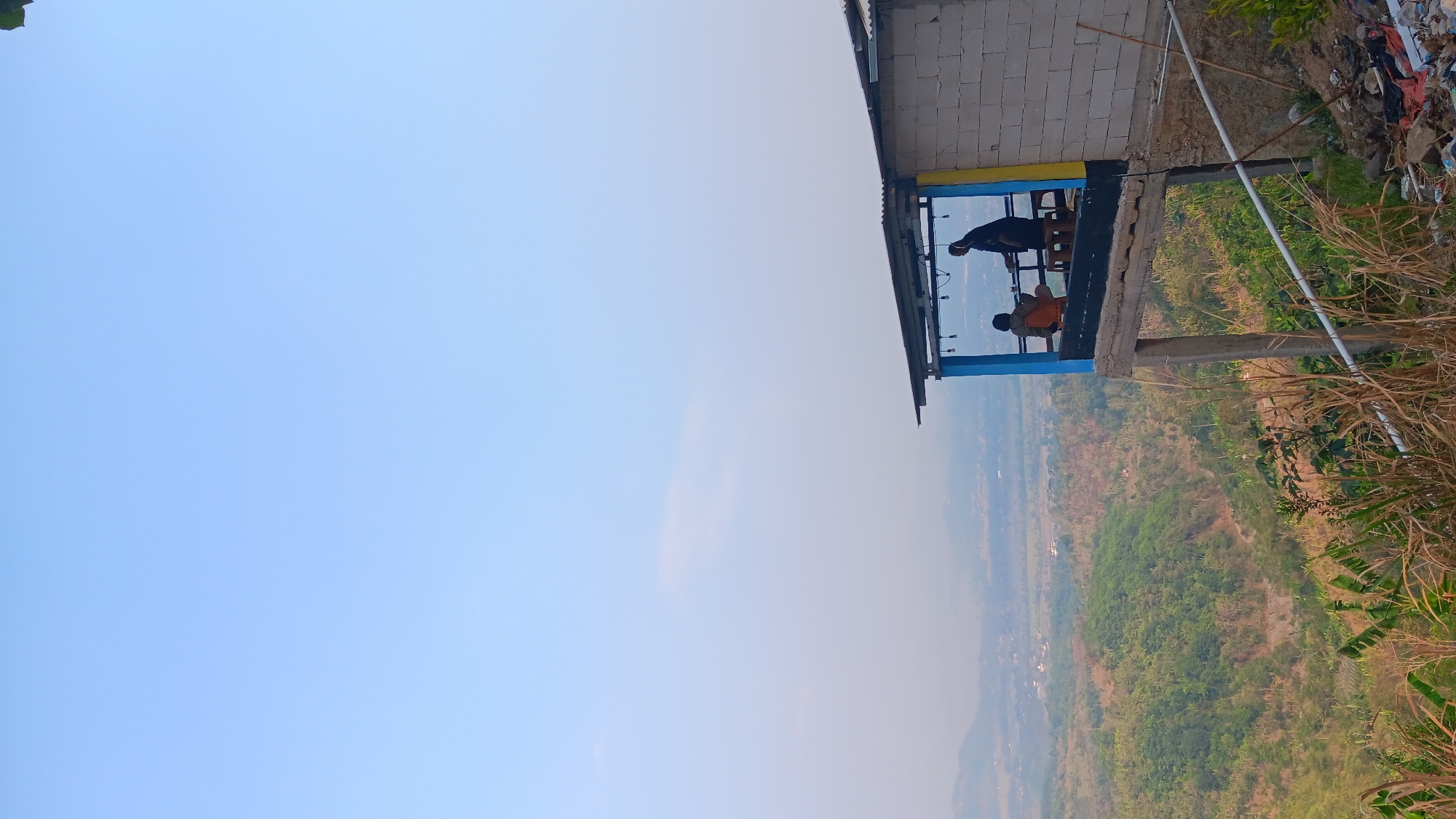The image depicts an elevated view of a rural landscape from a building's balcony. In the foreground, a small, open-air structure with a corrugated metal roof is visible, supported by concrete pillars. The structure's front is adorned with blue and yellow accents. Two individuals are present on the balcony: one person is seated at a table, and another is standing. The background reveals a vast expanse of rolling hills covered in lush green vegetation, interspersed with patches of dry earth and some scattered structures, suggesting a village or town within the valley. The sky above is a clear, pale blue, indicating fair weather. The scene is likely set in or around Mekargalih, Indonesia, given the geographical and architectural context commonly found in such areas. The overall impression is one of a serene, natural environment with a simple, functional human habitation offering a vantage point for observation.