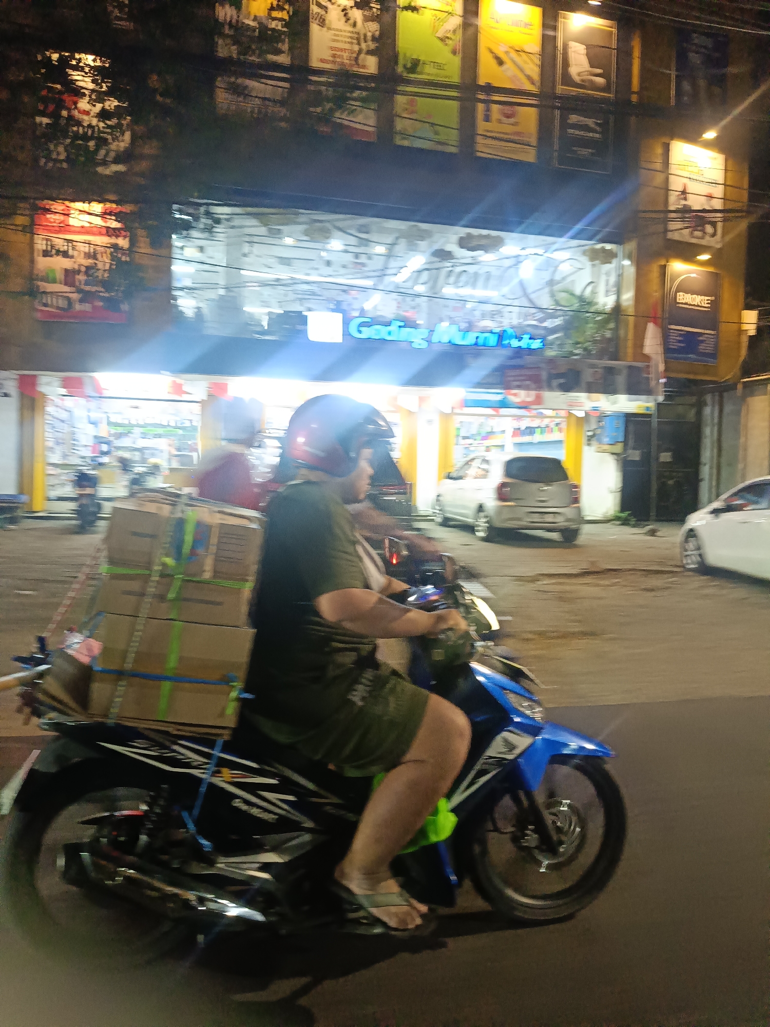 In Surabaya, Indonesia, a person wearing a red helmet and a dark green shirt is riding a blue motorcycle at night. The motorcycle is loaded with several cardboard boxes secured with green and blue straps. The boxes appear to be stacked high behind the rider. The background shows a lit-up building with various signs and posters, suggesting it is a commercial establishment. One sign, visible in blue neon letters, reads "Gading Murni Auto." A silver car is parked in front of the building, and another white car is visible on the right side of the image. The street appears to be paved. The overall scene depicts a nocturnal street view in a commercial area of Surabaya.