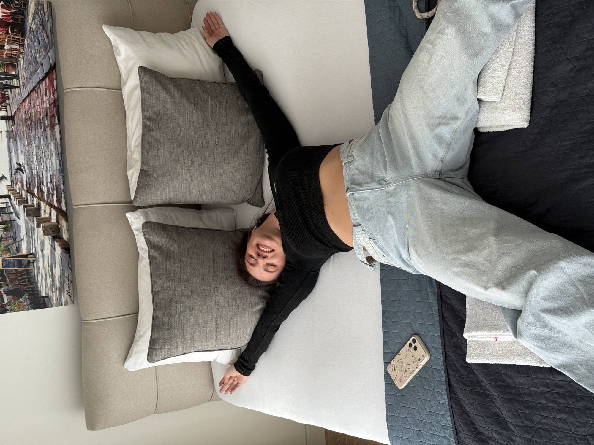 The image depicts an indoor scene, characteristic of a hotel or residential bedroom, located in Belgrade, Serbia. A single female individual is lying supine on a bed, centered in the frame. Her arms are extended laterally, hands open, resting on the white bedding. Her head is tilted slightly to her right (viewer's left), with her eyes closed and mouth open in a smile, indicating a relaxed or joyful state. She is dressed in a black long-sleeved cropped top, exposing her midriff, and light-wash blue denim jeans. Her dark hair is casually spread on the pillow area.
The bed features a light beige upholstered headboard against a plain light-colored wall. Two white pillows and two textured grey accent pillows are arranged against the headboard. The mattress is covered with a white fitted sheet. At the foot of the bed, a blue bed runner with a geometric pattern overlies a dark grey or black quilted comforter. To the left of the individual's torso, a light-colored smartphone with a floral-patterned case (pink flowers on a beige background) and a visible triple camera lens array is placed face-up on the blue bed runner. Two white folded towels are positioned on the dark comforter beneath the individual's left leg.
Above the headboard, a large landscape painting is hung, depicting an outdoor European street scene with cobblestones, pedestrians, buildings, and cafe seating. A signature, "Maracad," is visible on the artwork. The specific address or neighborhood within Belgrade is not discernable from visual cues in the image.
