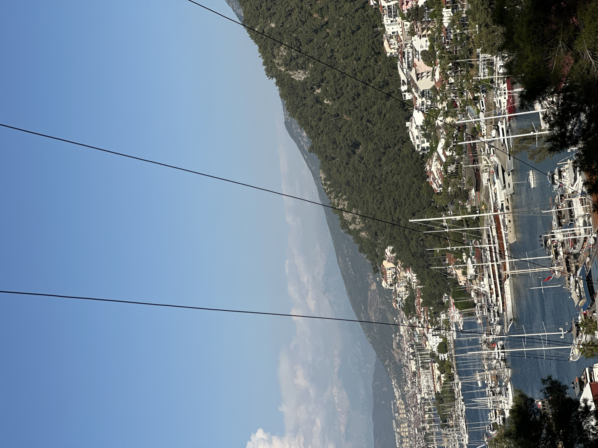 This panoramic image, when viewed in its correct orientation, captures a vibrant coastal scene, likely Fethiye, Turkey, from an elevated vantage point. The foreground is dominated by a bustling marina filled with numerous boats, primarily large white yachts and sailboats, their tall masts creating a dense pattern against the calm blue waters of the bay. Many boats are docked, suggesting a period of leisure or mooring.
Along the curved shoreline, a dense cluster of buildings with white walls and terracotta or reddish roofs forms the town. These structures extend from the waterfront and climb up the gentle slopes of the surrounding hills, which are thickly covered with lush green trees. In the background, majestic mountains rise, also heavily forested, and recede into a soft, atmospheric haze under a clear, bright blue sky. A few wispy clouds are visible in the upper sky.
The scene is bathed in strong, bright daylight, indicating late morning or early afternoon on a clear, sunny day. A series of dark, thin power lines stretch across the upper portion of the frame, suggesting the viewpoint is from a cable car or a high location near infrastructure. No discernible text is visible within the image. The overall impression is one of a picturesque and active Mediterranean port town nestled between verdant mountains and the sea.