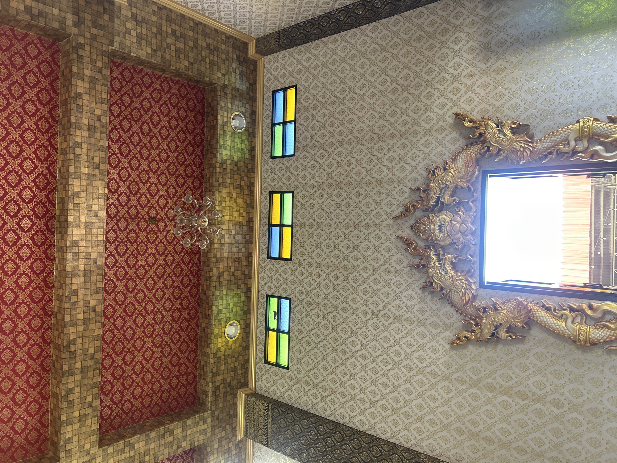 The image shows the interior of a building with ornate decorations. The ceiling is a prominent feature, with a red fabric adorned with a gold diamond pattern, framed by a textured gold border. Below the ceiling, there are three rectangular stained-glass windows with colorful panes in yellow, blue, and green. The walls are covered in a light-colored wallpaper with a subtle gold damask pattern. In the foreground, a doorway is framed by elaborate golden dragon sculptures, their heads facing outwards. The doorway leads to a brightly lit area, suggesting an exit or another part of the building. The overall impression is one of richness and traditional artistry. This scene is located in the Thai Ko Subdistrict, Thailand, likely within a temple or a similarly culturally significant structure. There are no people visible in the image.