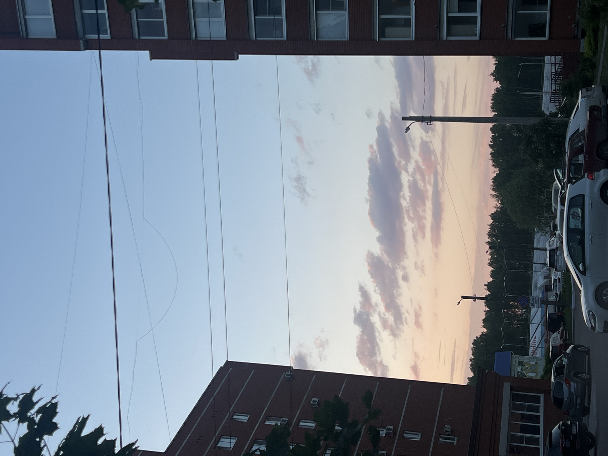 The image shows a scene in Chaikovsky, Russia, during what appears to be a sunset. Two brick buildings frame the view of the sky, with power lines running across the open space. The sky is a mix of light blue and pinkish-orange hues, with elongated clouds. In the distance, a row of trees and parked cars can be seen. One of the buildings has numerous windows, and there are some trees visible at the bottom of the image. There are lampposts visible near the trees and parked cars.