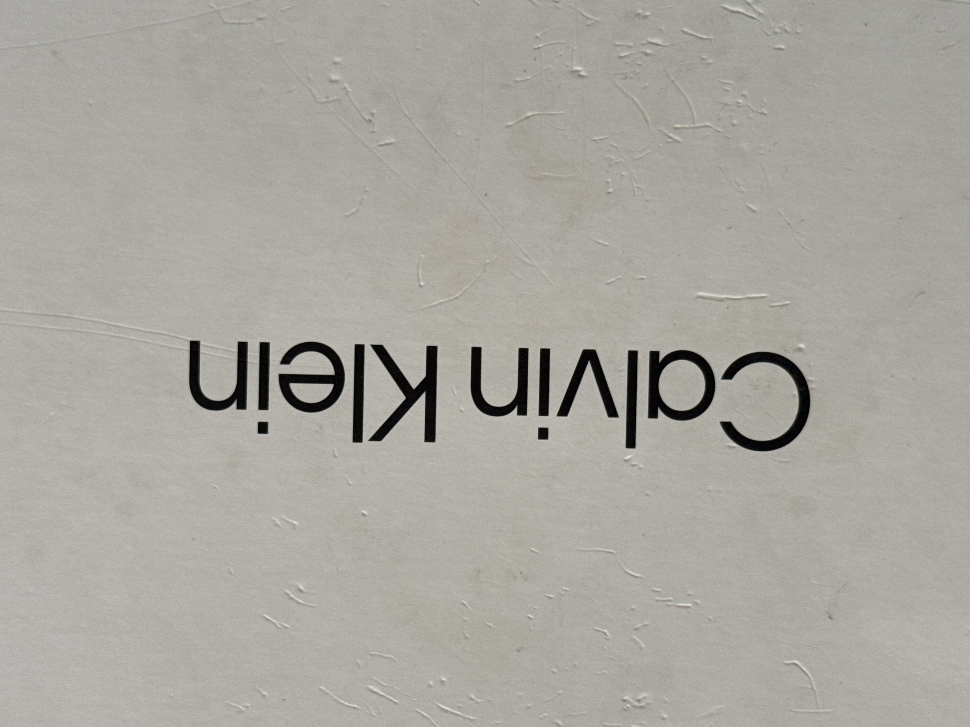 The image displays the brand name "Calvin Klein" printed vertically in black, sans-serif capital letters against a textured, light grey background. The background surface exhibits irregular raised lines, shallow indentations, and minor discolorations, consistent with a rough or worn material such as plaster or canvas. The text is oriented from bottom to top. No human subjects, actions, or specific interactions are visible within the frame. This scene is located in Busmantsi, Bulgaria.