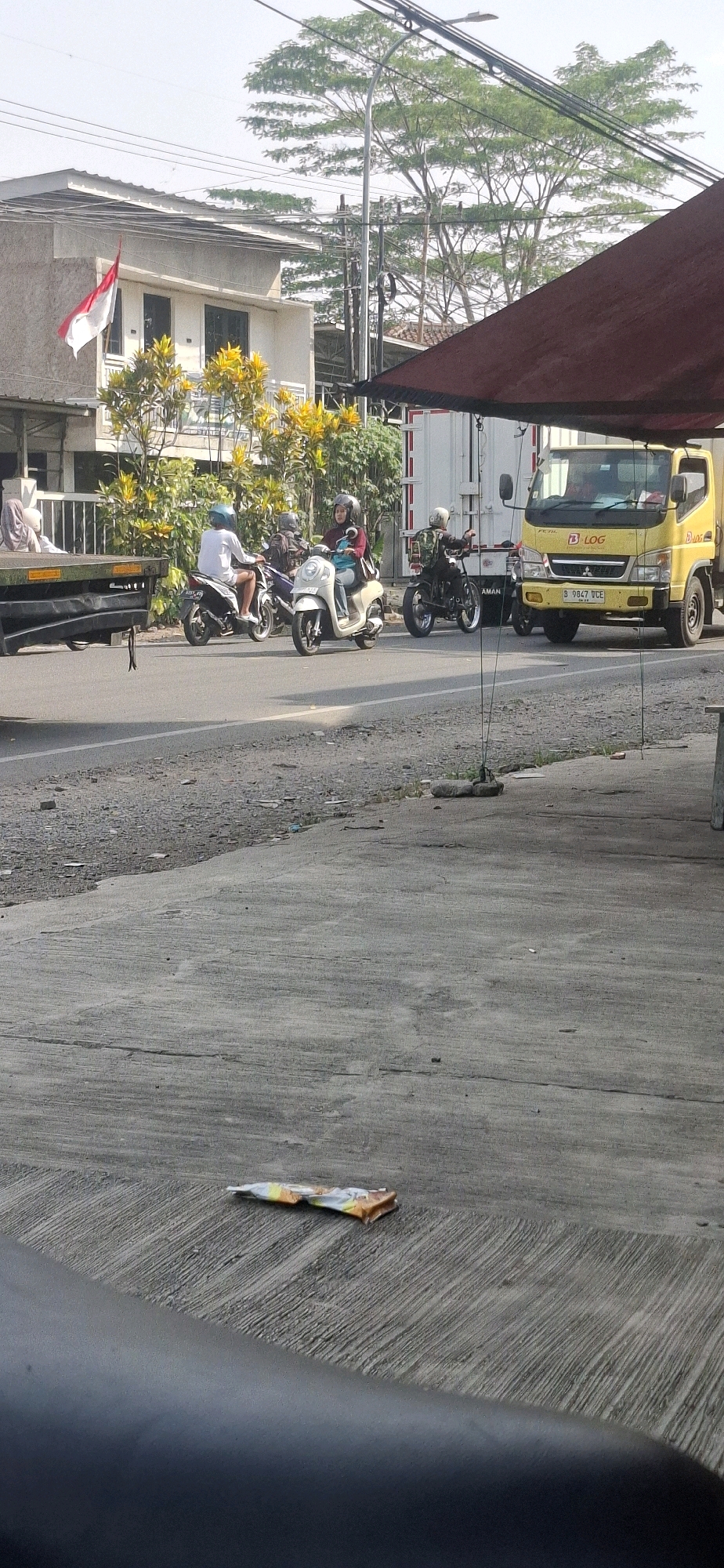 In the city of Rancagoong, Indonesia, on a sunny day, several motorcycles are visible on the road. One motorcycle, a white automatic scooter, carries a female rider wearing a blue shirt and a helmet, and a passenger in a dark hijab and a maroon top, also wearing a helmet. Another motorcycle, a dark scooter, has a rider in a white shirt and shorts, wearing a light blue helmet. To the right of these, a third motorcycle, a black model, has a rider wearing a backpack and a helmet. A yellow truck with the logo "LOG" on its side and a license plate that reads "B 9847 VCE" is parked on the right side of the road, partially obscured by a red awning. In the background, a two-story building with an Indonesian flag displayed on its upper floor is visible, surrounded by trees with yellow foliage. A person is lying down on a flatbed trailer on the left side of the image. The foreground shows a concrete surface with some gravel and debris, and a crumpled food wrapper is lying on the concrete. Power lines are visible stretching across the sky.