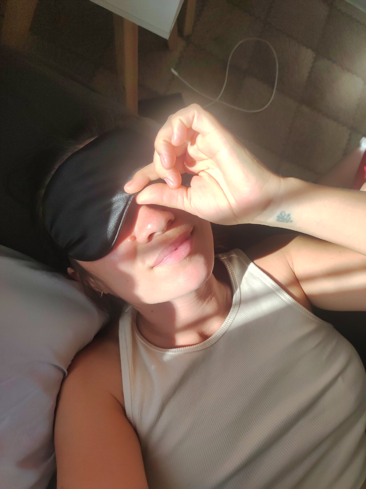 A woman is lying down wearing a black sleep mask and a white ribbed tank top. Her right hand is raised, holding the sleep mask over her eyes. Sunlight is casting shadows on her face and chest. A white cord is visible on the floor behind her. The scene appears to be indoors, possibly in a bedroom, with a dark surface and a floor covered in light-colored tiles or a similar material. There is a pillow on the left side of the image. There is a small tattoo on her wrist.