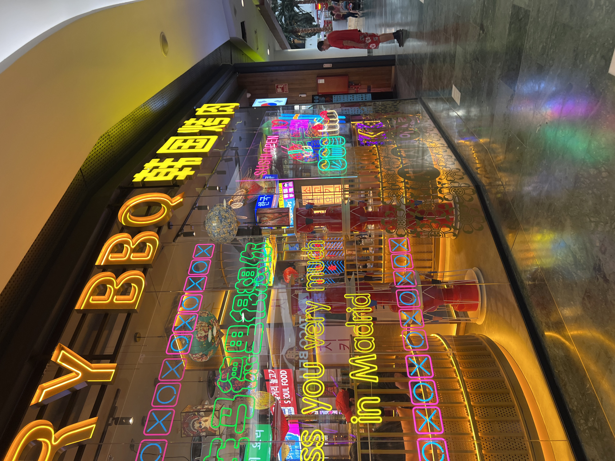 The image captures the exterior of a brightly lit Korean BBQ restaurant in Madrid, Spain. The restaurant's facade is dominated by neon signage, with large yellow letters spelling out "RY BBQ" and Korean characters. Below this, various neon signs in pink, blue, and green display playful designs, including tic-tac-toe grids and what appears to be text in Korean and English, such as "SOUL FOOD" and "SUSHI OPEN." A prominent neon sign also reads "ss you very much in Madrid."
Inside the restaurant, visible through the glass, are decorative elements like hanging lanterns and what seem to be red spinning structures. A man wearing a red t-shirt and patterned shorts stands outside the restaurant, facing away from the camera and looking towards the entrance. In the background, other individuals and palm trees suggest an indoor shopping mall environment. The floor is made of dark, polished tiles that reflect the colorful neon lights. The overall impression is a vibrant and modern establishment, likely popular for its visual appeal and diverse offerings.