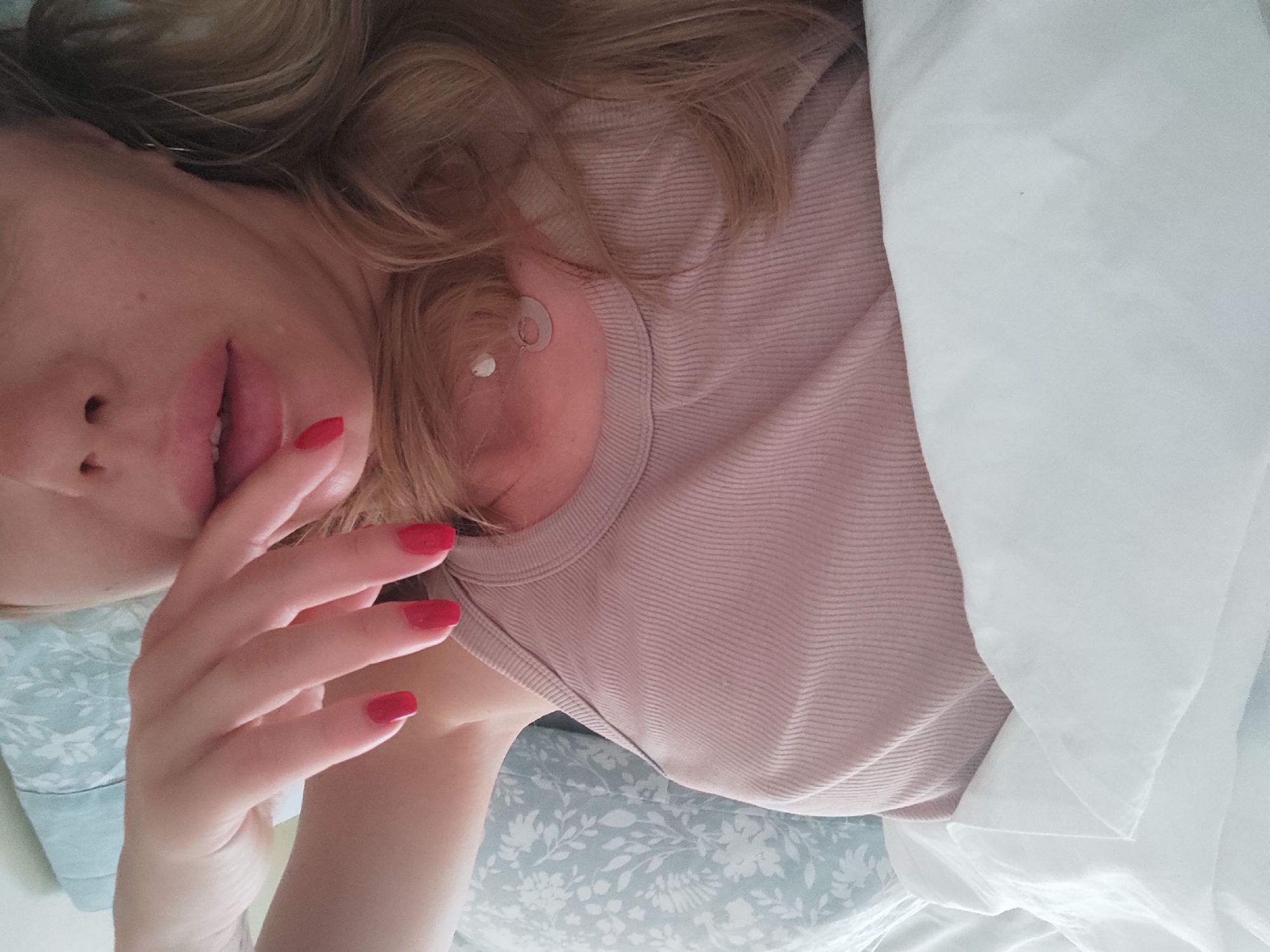 A person is lying on a bed with white and blue floral patterned sheets. The person is wearing a light pink ribbed tank top. Their right hand, with red nail polish, is raised towards their face, with the index finger touching their lips. Their mouth is slightly open, and their tongue is visible. The person has blonde hair that is spread around their head. A silver earring with a light blue stone is visible on their left ear. The overall scene suggests a relaxed or intimate setting, possibly a bedroom. There is no specific identifiable location within the city of Blanes, Spain.