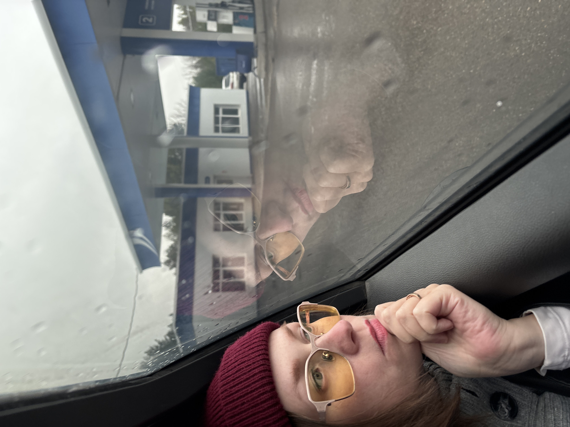 The image shows a woman wearing glasses and a red beanie, looking out of a car window on a rainy day. Her reflection is visible in the wet glass. Outside the window is a gas station. The specific location cannot be definitively determined as the details available are insufficient.