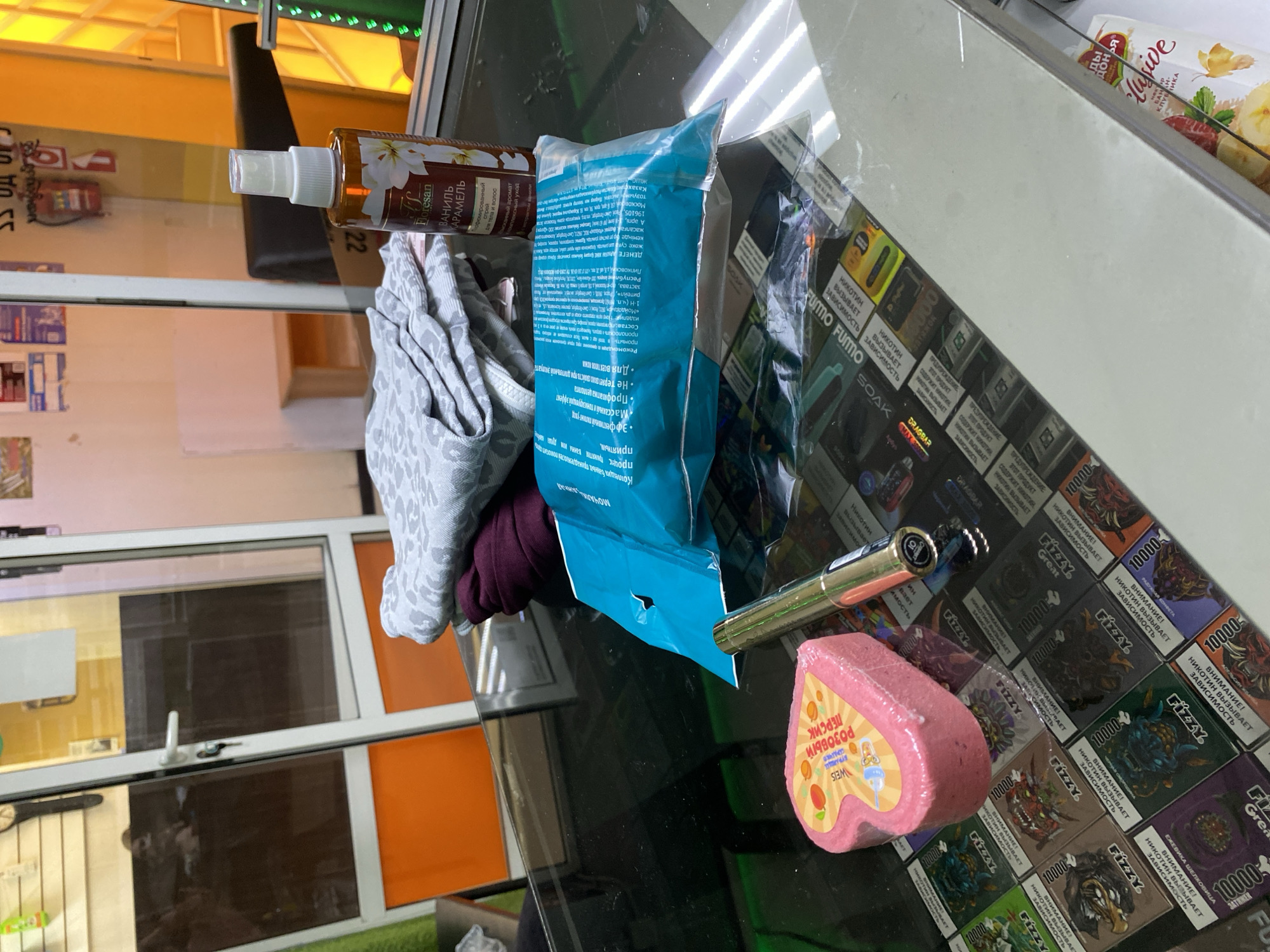 The image displays various retail products arranged on a reflective glass surface, likely a store counter. No people are visible.
Prominent objects include a brown spray bottle labeled "Floresan ВАНИЛЬ КАРАМЕЛЬ" (Floresan Vanilla Caramel) described as a perfumed spray for body and hair. Next to it is a blue plastic bag, partially obscuring a white and grey patterned fabric, possibly a folded towel or garment. The bag's label includes "МОЧАЛКИ" (Washcloths) and extensive Russian text detailing product features like "Для всех типов кожи" (For all skin types) and references to "Казахстан" and "Москов".
Further right, a gold-colored pen-like object lies near a heart-shaped pink bath bomb labeled "WEIS РОЗОВЫЙ ПЕРСИК СЕРДЕЧКО" (WEIS Rosy Peach Heart). Beneath these items, and covering a significant part of the counter, are numerous boxes of electronic cigarettes or vape devices from brands like "Fizzy," "Fumo," "Dragbar," and "Soak." Many of these boxes prominently display warnings in Russian: "НИКОТИН ВЫЗЫВАЕТ ЗАВИСИМОСТЬ" (NICOTINE CAUSES ADDICTION) and "ПРЕДУПРЕЖДЕНИЕ: ЭТОТ ПРОДУКТ СОДЕРЖИТ НИКОТИН" (WARNING: THIS PRODUCT CONTAINS NICOTINE). Some boxes indicate "10000 затяжек" (10000 puffs) and flavor names such as "ЕЖЕВИКА ШЕЛКОВИЦА" (Blackberry Mulberry) and "КЛУБНИКА" (Strawberry).
The glass surface reflects an indoor environment with large windows or glass panels and orange-brown wall sections, typical of a retail space. The visible text is primarily in Russian, suggesting the location is in a Russian-speaking region. The time of day and weather are not discernible from the indoor setting.