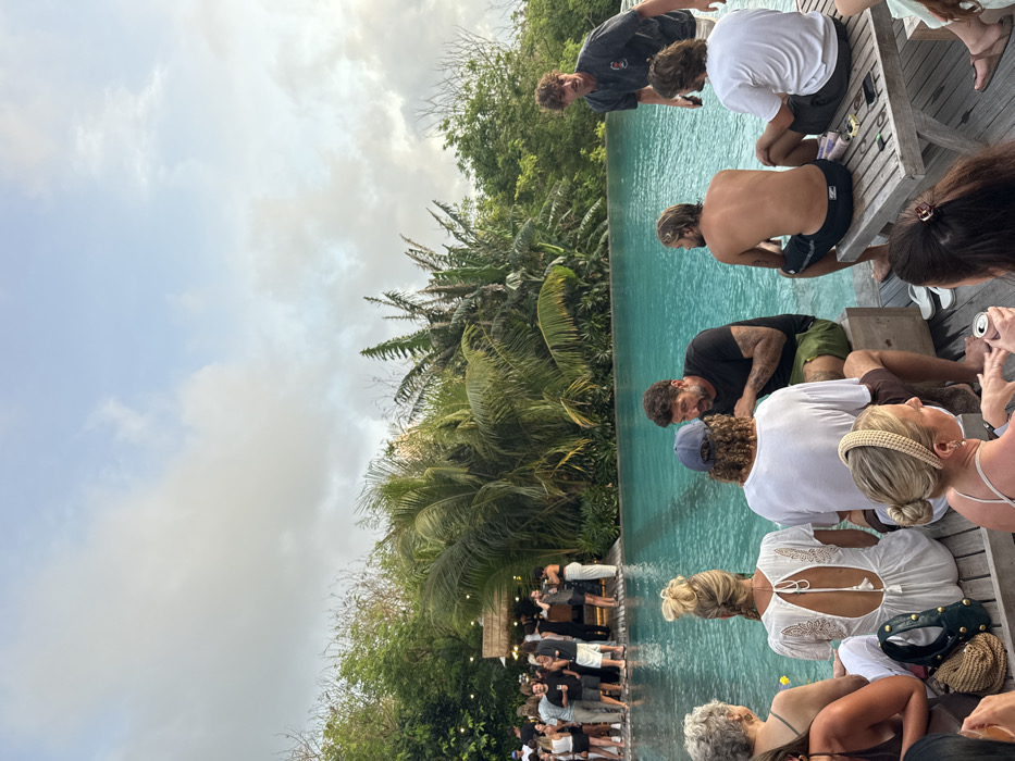 This image depicts an outdoor social gathering taking place at an establishment in Uluwatu, Indonesia, likely a resort or beach club, centered around a turquoise infinity pool framed by dense tropical vegetation under a partly cloudy sky during what appears to be late afternoon or early evening.
In the immediate foreground, a group of approximately five individuals is seated on a wooden deck, partially facing away from the camera. From left to right, one individual with short grey hair is seen from behind, followed by a person with blonde hair in a bun wearing a white open-backed top with embroidered details. Next, an individual wearing a blue baseball cap and white t-shirt is seated on a light wooden block, conversing with a bearded man in a black t-shirt and olive-green shorts, who is also seated on a wooden block. The bearded man has visible tattoos on his left arm. Further right, a shirtless man with long blonde hair is seated on a wooden bench, wearing dark swim shorts, and next to him, another man with dark, longish hair wearing a white t-shirt is also seated on the bench. On the far right foreground, a portion of a wooden table is visible with white flip-flops, a black smartphone, and several small items on its surface.
In the midground, the large, clear blue infinity pool dominates, reflecting the sky. On the opposite side of the pool in the background, a cluster of approximately two dozen individuals is gathered around a rustic-style bar or hut, constructed from natural materials and adorned with string lights. These individuals are standing, conversing, and holding drinks. The entire background beyond the pool and bar is filled with lush, vibrant green tropical foliage, including large palm trees and other broad-leafed plants, creating a dense natural wall. The sky above is a mix of light blue and soft grey clouds. The overall scene conveys a relaxed, social atmosphere.