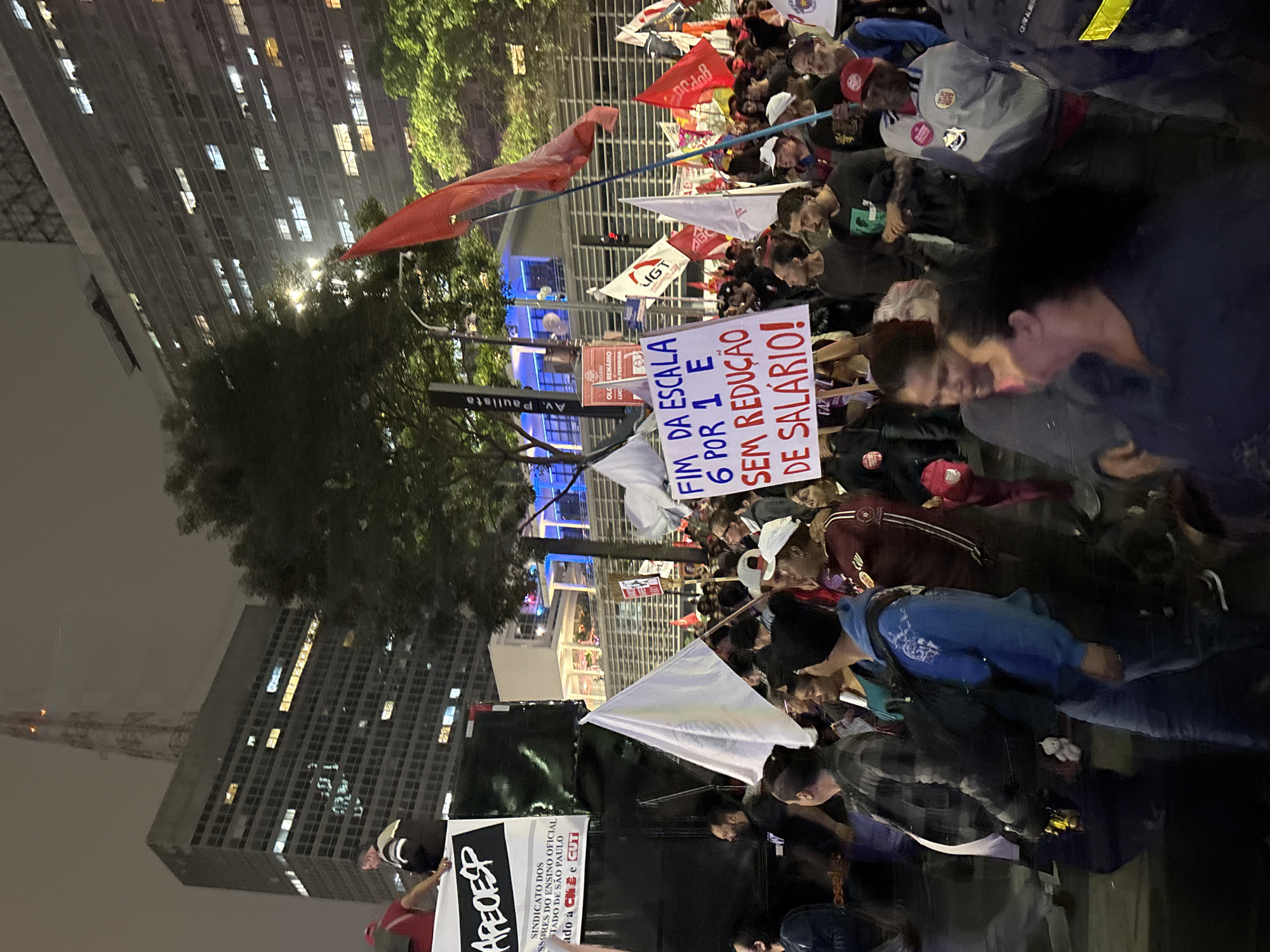A nighttime image depicts a crowd of people, likely a demonstration or protest, on Avenida Paulista in São Paulo, Brazil. APECESP and other signs are visible in Portuguese. The sign reads "FIM DA ESCALA 6 POR 1 E SEM REDUÇÃO DE SALÁRIO!" ("END OF THE SCALE 6 FOR 1 AND NO SALARY REDUCTION!")
The setting features urban architecture, including tall buildings. Some buildings have their windows lit up, indicating occupancy. The crowd consists of a diverse group of people. There are red and white flags. The overall atmosphere suggests a political gathering.