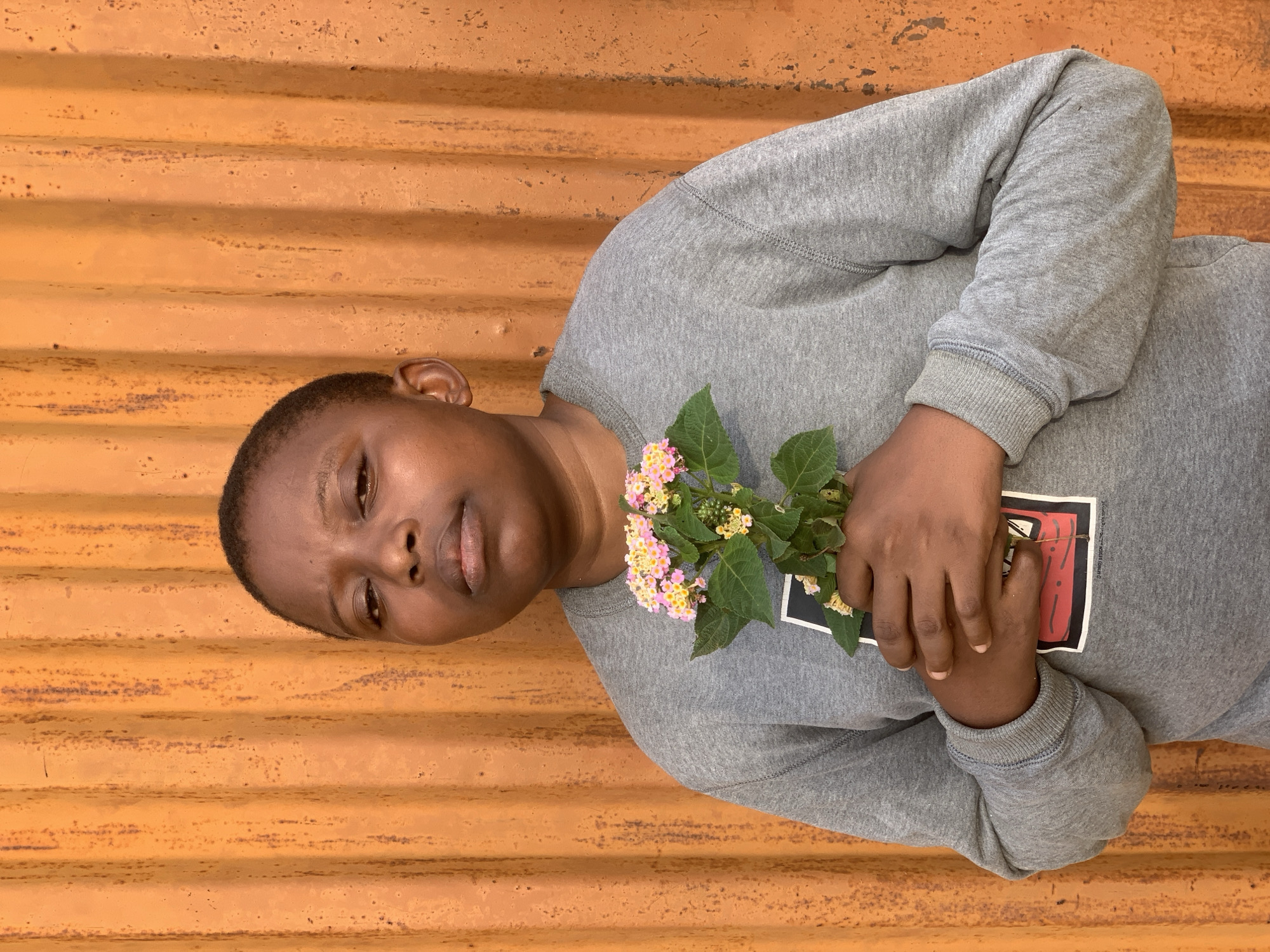A person lies angled against an orange corrugated metal wall. The person, wearing a gray sweatshirt with a design on the chest, holds a small bouquet of yellow and pink flowers in their hands. The location is presumed to be Ikorodu, Nigeria.