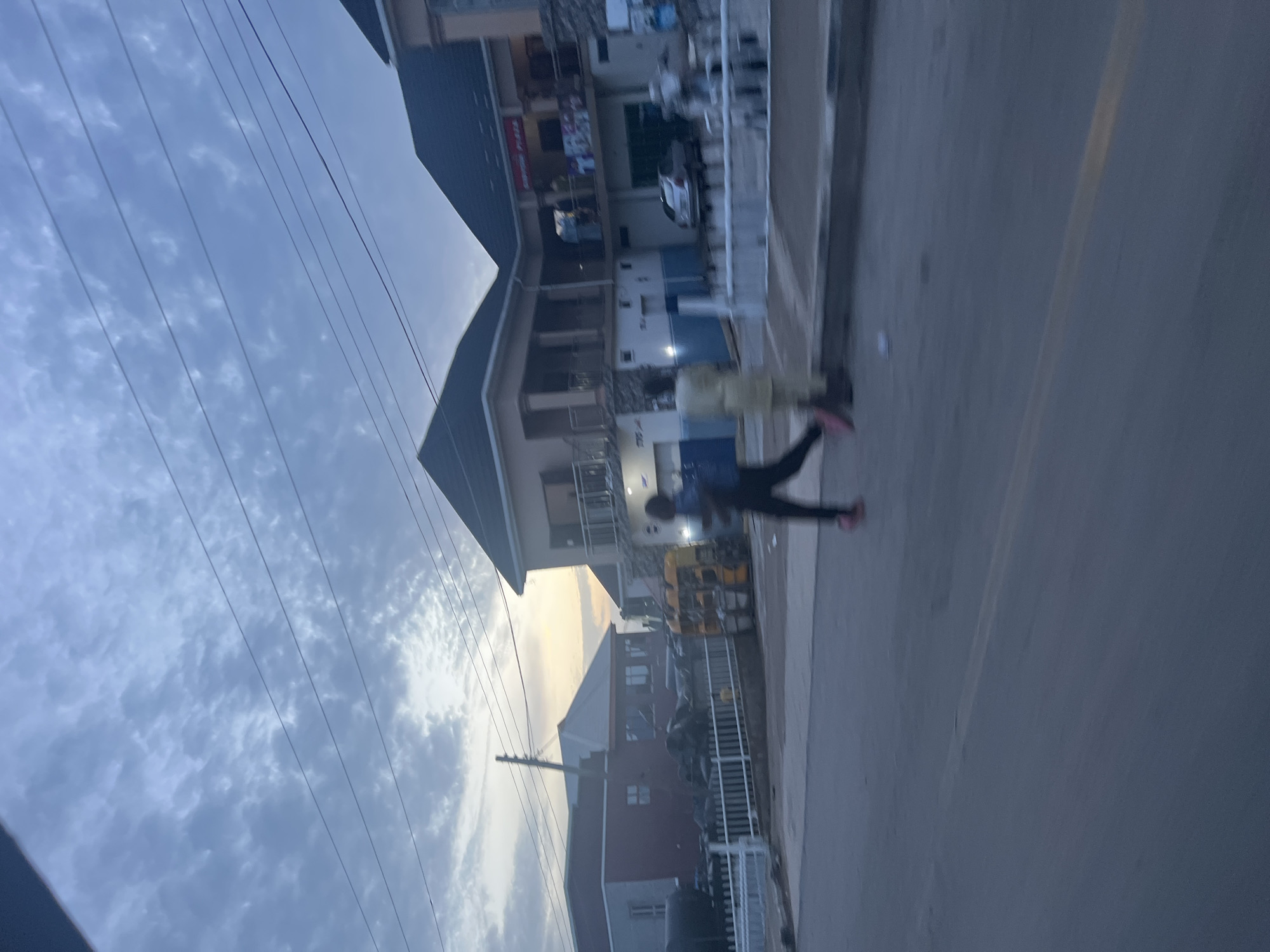The image depicts a street scene in Jalingo, Nigeria, under a cloudy sky. In the foreground, a paved road with a faint yellow line is visible. Two individuals are in motion. One person, wearing dark clothing and pink footwear, is running across the street, appearing to be in mid-stride. Another individual, dressed in light-colored traditional attire, stands further away on the sidewalk.
Behind them, a multi-story building with a dark roof and balconies stands prominently. The building has an advertisement for "Shuon Plaza" on its facade. Below the plaza, a covered area appears to be a garage or shopfront, with a silver car parked inside. To the left of this building, another, lower structure with a red roof is partially visible. Further to the left, a white fence runs along the edge of a property. A yellow auto-rickshaw is parked near the red-roofed building. Overhead, multiple power lines crisscross the sky. The overall scene suggests a typical urban environment in Jalingo during the early morning or late afternoon, judging by the soft light.