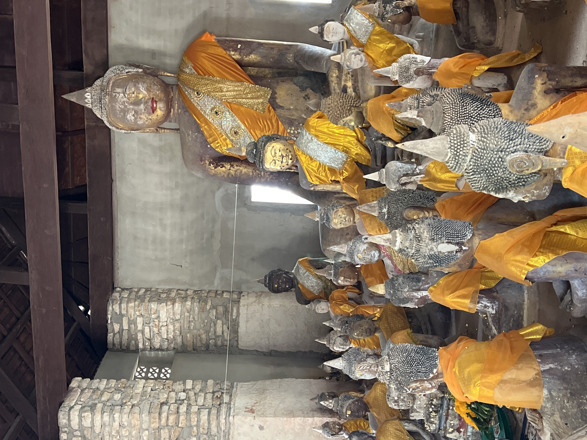 The image depicts a collection of Buddha statues inside a building in Baan Harn, Thailand. The main subject is a large, seated Buddha statue at the rear, draped in an orange and gold robe. In front of it, and surrounding it, are numerous smaller Buddha statues, many also adorned with orange fabric. The statues vary in size, material, and detail, with some appearing weathered and antique. The interior environment features rough, light-colored stone walls and a wooden roof structure with visible beams. Sunlight streams in from the right, illuminating parts of the scene. There are no people visible in the image.
