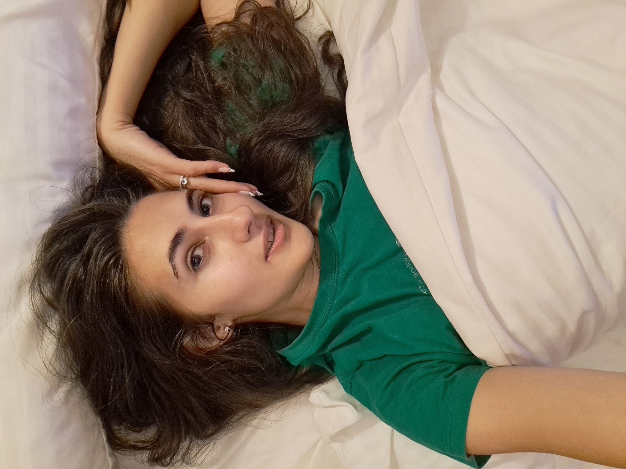 A woman with dark hair is lying in a bed covered with a white sheet and pillow. She is wearing a green shirt. Her left hand is raised to her forehead, and she is wearing a ring on her finger. The background is a white bedsheet. The location of Negovan, Bulgaria is not discernible in the image.