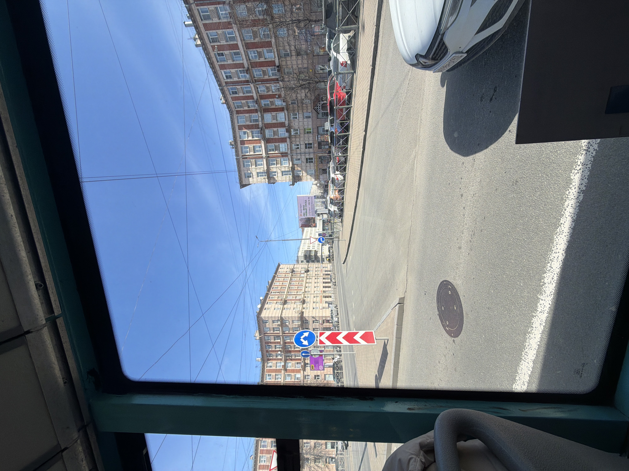 The view from inside a vehicle, possibly a bus or tram, showcases a street scene in Saint Petersburg, Russia. The street is lined with multi-story residential buildings, predominantly brick and white stone. The sky is a clear blue. A row of parked cars is visible on the right side of the street. A road sign indicating a sharp turn with a chevron pattern is prominently displayed. Other street signs and a public advertisement are also visible. The roadway is asphalt. The vehicle's window frame is green.