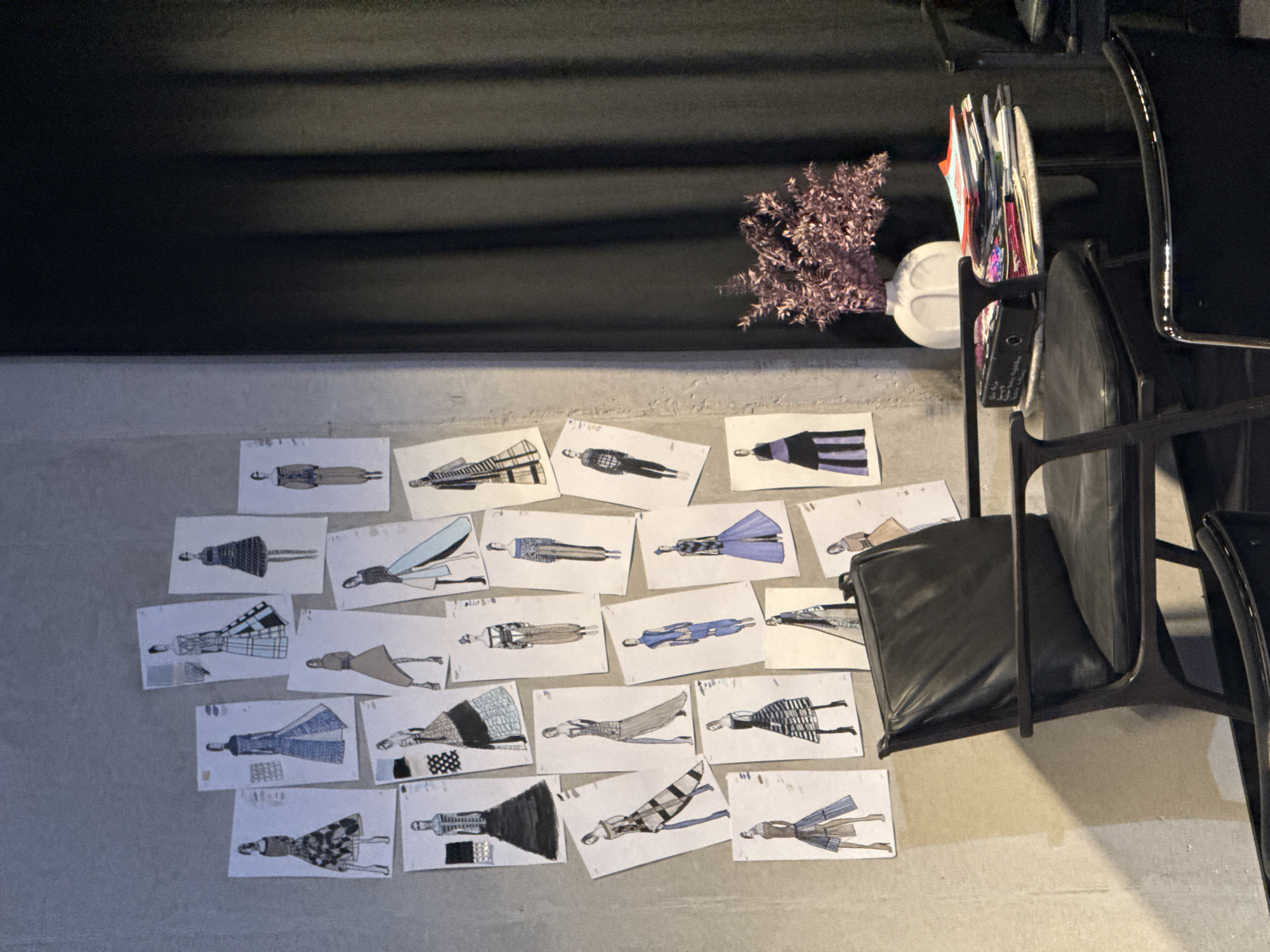 An interior scene in Sofia, Bulgaria, features a light-grey wall on the left and center, densely covered with approximately 30-35 individual fashion design sketches. These sketches, rendered on white paper, depict female figures in various poses, wearing diverse outfits that exhibit patterns, textures, and a color palette including blues, blacks, browns, purples, and light greens. In the foreground, a dark, possibly leather-upholstered armchair with dark wooden arms is centrally positioned. To its immediate right, a small, round table holds a stack of colorful magazines or books, a black binder, and a white, sculpted vase containing dried purple foliage. The right side of the frame is dominated by a dark, vertically pleated curtain or fabric panel. Partially visible in the lower right are sections of other dark chairs, one of which features a chrome trim.