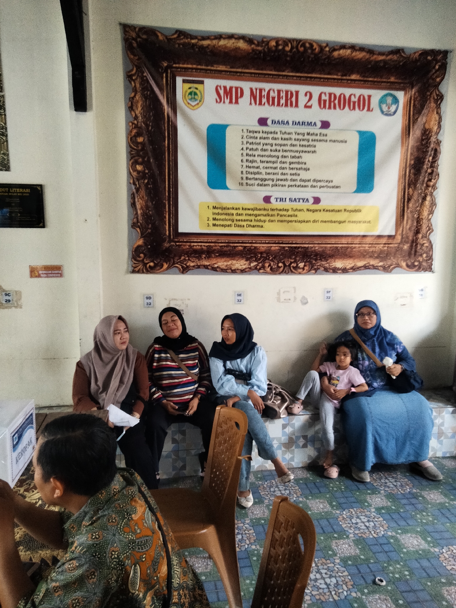 In Sanggrahan, Indonesia, a group of people, primarily women, are gathered in what appears to be an indoor public space. Dominating the background is a large framed banner for "SMP NEGERI 2 GROGOL," an Indonesian junior high school, displaying the school's emblem and mottos. One woman in a blue hijab and a long blue skirt is seated on a tiered bench, holding a small child. To her left, another woman in a blue hijab and a light blue top is seated, while to her right, two women are seated, one in a striped shirt and a headscarf, and another in a light brown hijab. In the foreground, a man wearing a patterned batik shirt is visible from the back, looking at a box labeled "KOPAK." The environment is brightly lit, with a patterned tiled floor and several brown plastic chairs. The overall scene suggests a community gathering or an administrative event at the school.