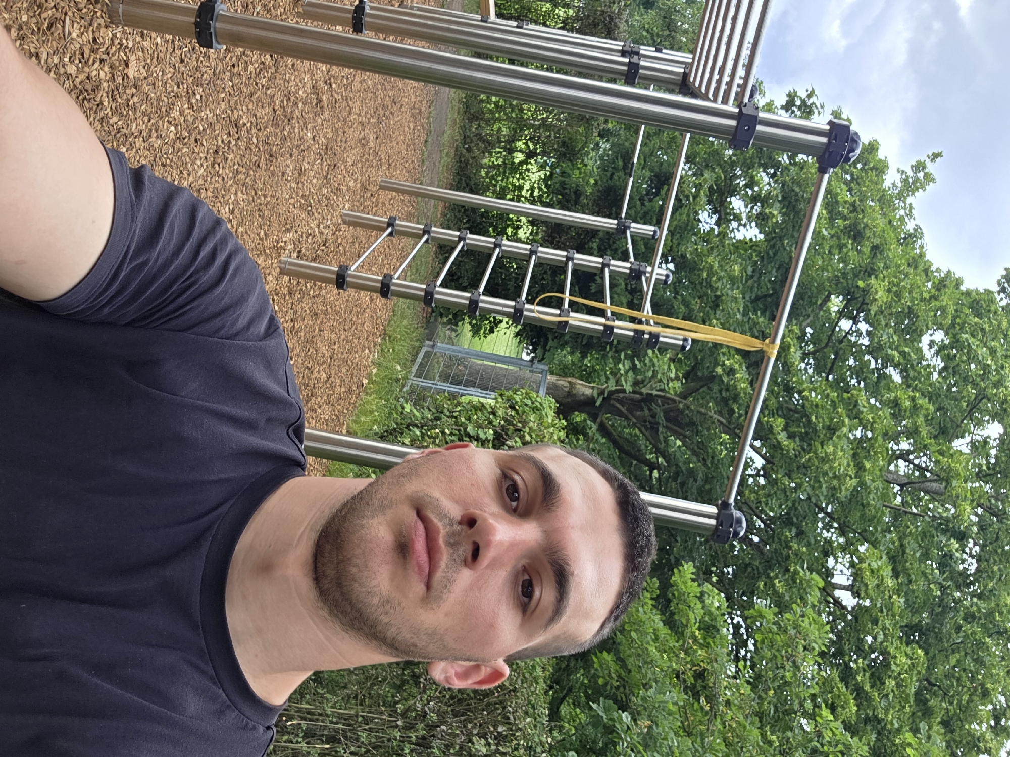 A young adult male with short dark hair and stubble is taking a selfie outdoors at a park. He is wearing a dark t-shirt and is positioned in the foreground, with a playground structure made of metal bars and a yellow resistance band visible behind him. The ground is covered with wood chips, and lush green trees and bushes form the backdrop under a cloudy sky. The park appears to be located in a suburban or rural area, as suggested by the natural surroundings and the absence of urban infrastructure.