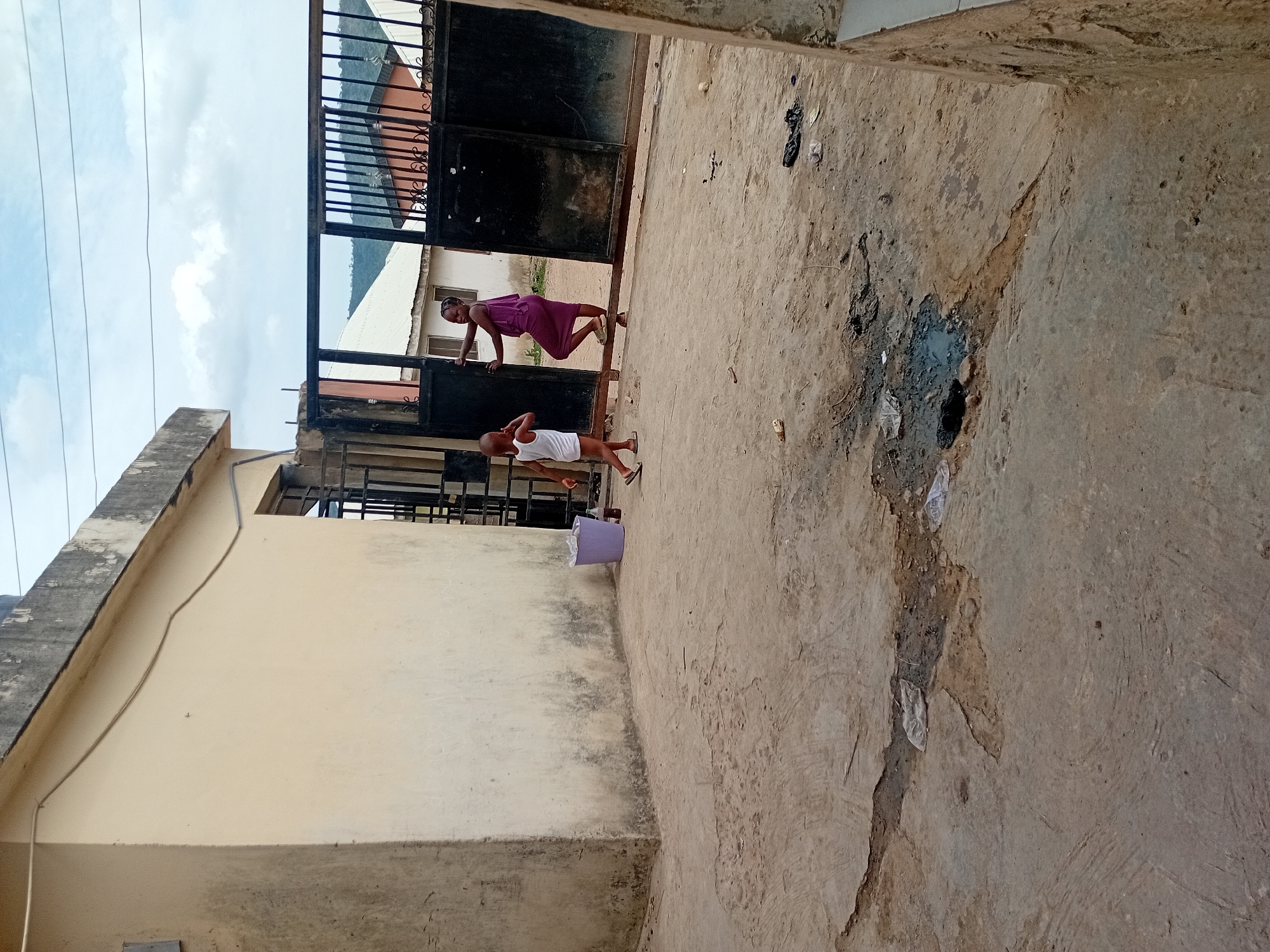 A medium shot depicts a weathered courtyard in an undetermined urban location. Two individuals are present: an adult woman in a purple dress climbs over a black metal gate, while a young boy in a white tank top and dark sandals stands near her. The ground is cracked and stained, with scattered debris. Adjacent to the gate is a beige building. Overcast sky visible above.
