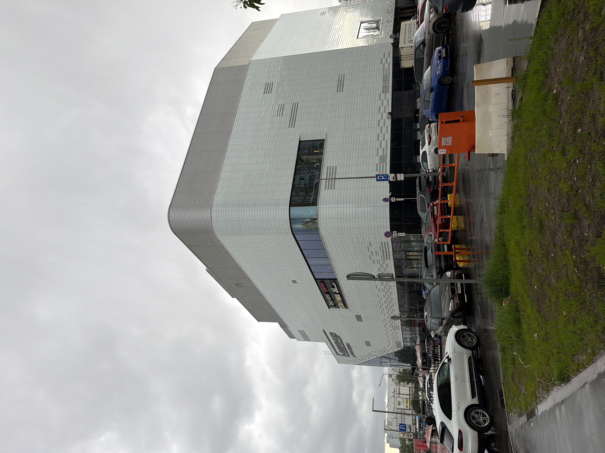 The image captures a view of the Okeania Shopping Mall in Moscow, Russia, on a cloudy and possibly wet day. The building's facade is predominantly white with a curved corner and horizontal lines, punctuated by rectangular windows and digital signage. Cars of various colors, including white, blue, black, and red, are parked along the street in front of the mall. Orange safety barriers and what appear to be concrete blocks are also present near the curb. The environment includes a narrow strip of green grass and a wet road surface, suggesting it has recently rained or is currently raining. The sky is overcast, casting a diffused light over the scene.