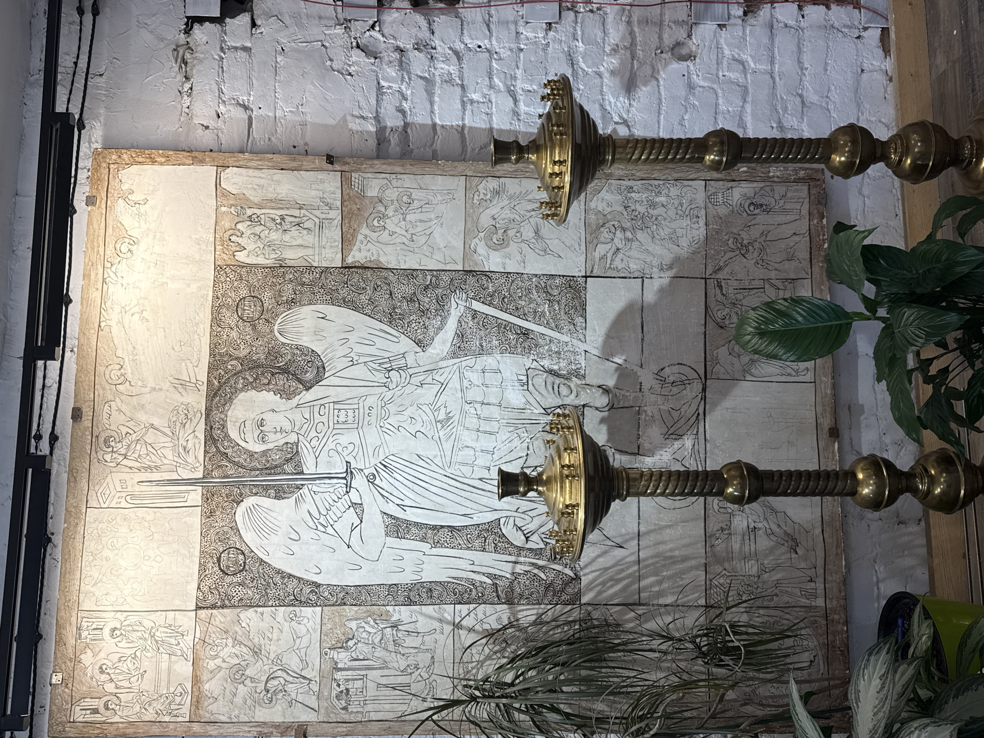 The image depicts an interior scene featuring a large, detailed drawing or painting on a canvas or parchment material, divided into a grid of square panels. The central and largest panel shows a winged figure, likely an angel, holding a sword and wearing armor. This central figure is surrounded by smaller panels, each containing individual scenes rendered in a similar line-art style, many of which appear to depict biblical or religious narratives with multiple figures. The artwork is mounted on a white brick wall. In the foreground, two ornate brass candelabra stand on a wooden surface. Lush green plants, including a tall, leafy plant on the left and a broad-leafed plant in the center, are also present in the foreground, adding a touch of nature to the setting. The overall ambiance suggests a spiritual or artistic space, possibly a church, chapel, or an art studio. The lighting is warm, illuminating the artwork and creating shadows. The image does not provide any visual clues to indicate the location as Yekaterinburg, Russia.