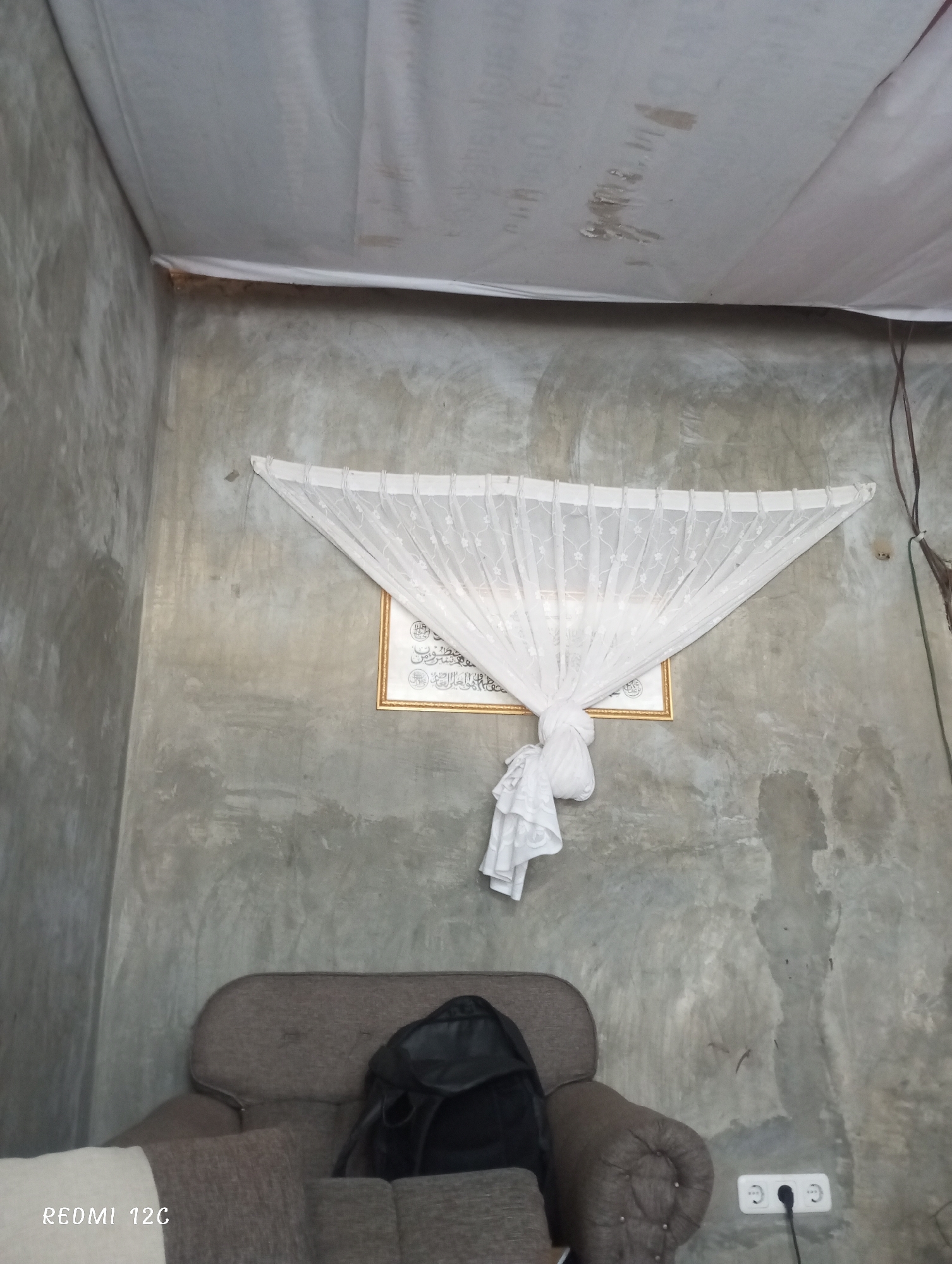Indoor scene in Lhokseumawe, Indonesia. Gray-toned room with textured walls and ceiling partially covered by a white sheet. A decorative white curtain hangs against the wall above a brown armchair. A black backpack is on the chair. A framed picture is behind the curtain. An electrical outlet is visible on the lower right wall. A sofa is visible on the left. The bottom left corner has "REDMI 12C" printed on it.